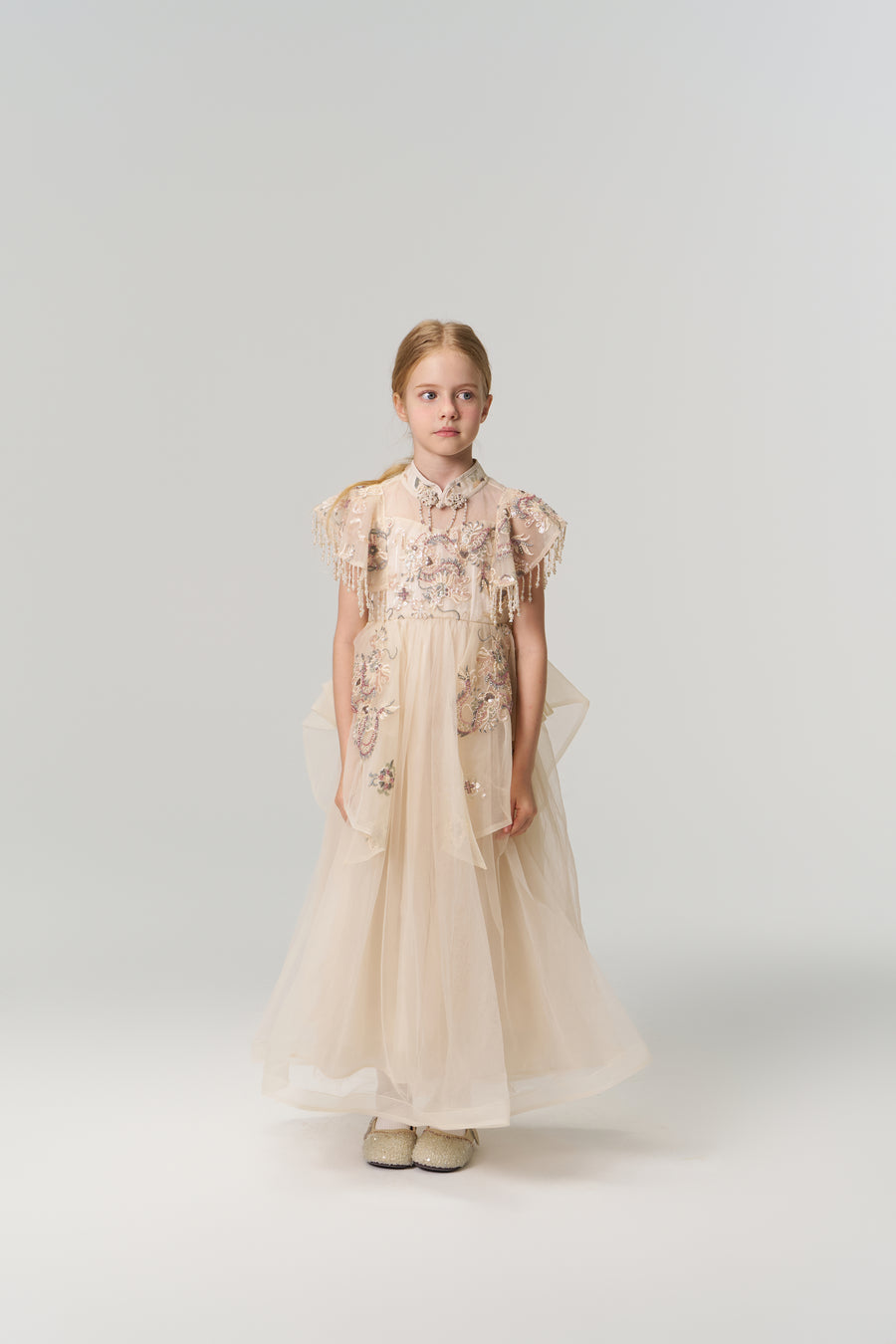 A young girl poses against a plain backdrop in the KIKI and JOJO Jade Whisper Dress, a long, light-hued cheongsam featuring embroidered florals, decorative sleeves, mandarin collar, and fluffy shoes.
