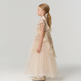 A young girl with long blonde hair stands in profile, wearing the ornate Jade Whisper Dress by KIKI and JOJO, featuring layered cream tulle, embroidered florals, and decorative embellishments against a plain light background.