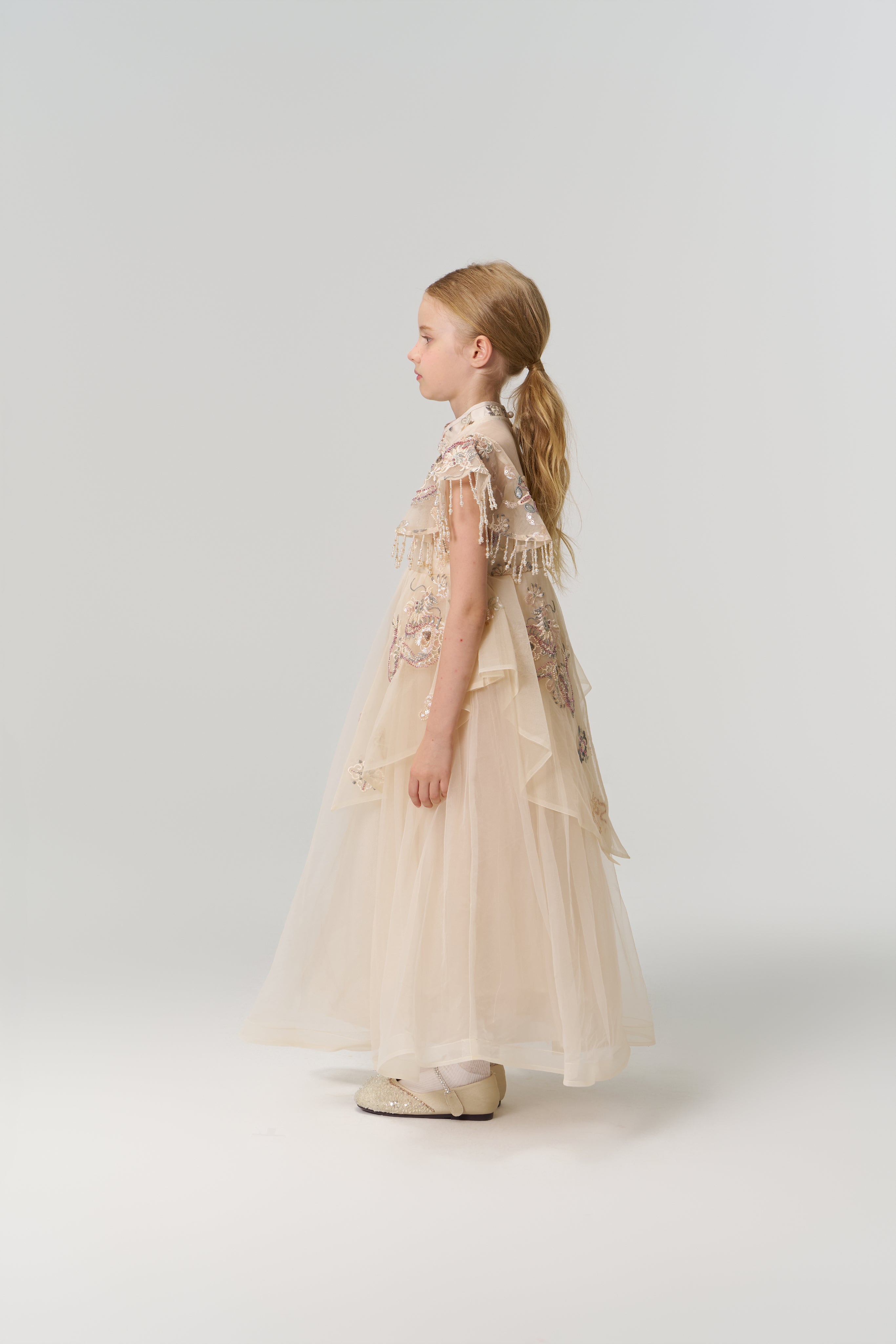 A young girl with long blonde hair stands in profile, wearing the ornate Jade Whisper Dress by KIKI and JOJO, featuring layered cream tulle, embroidered florals, and decorative embellishments against a plain light background.