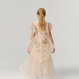 A young girl with blonde hair in a ponytail stands facing away, modeling the KIKI and JOJO Jade Whisper Dress—a long, light pink cheongsam with embroidered florals and a mandarin collar—against a plain white background.
