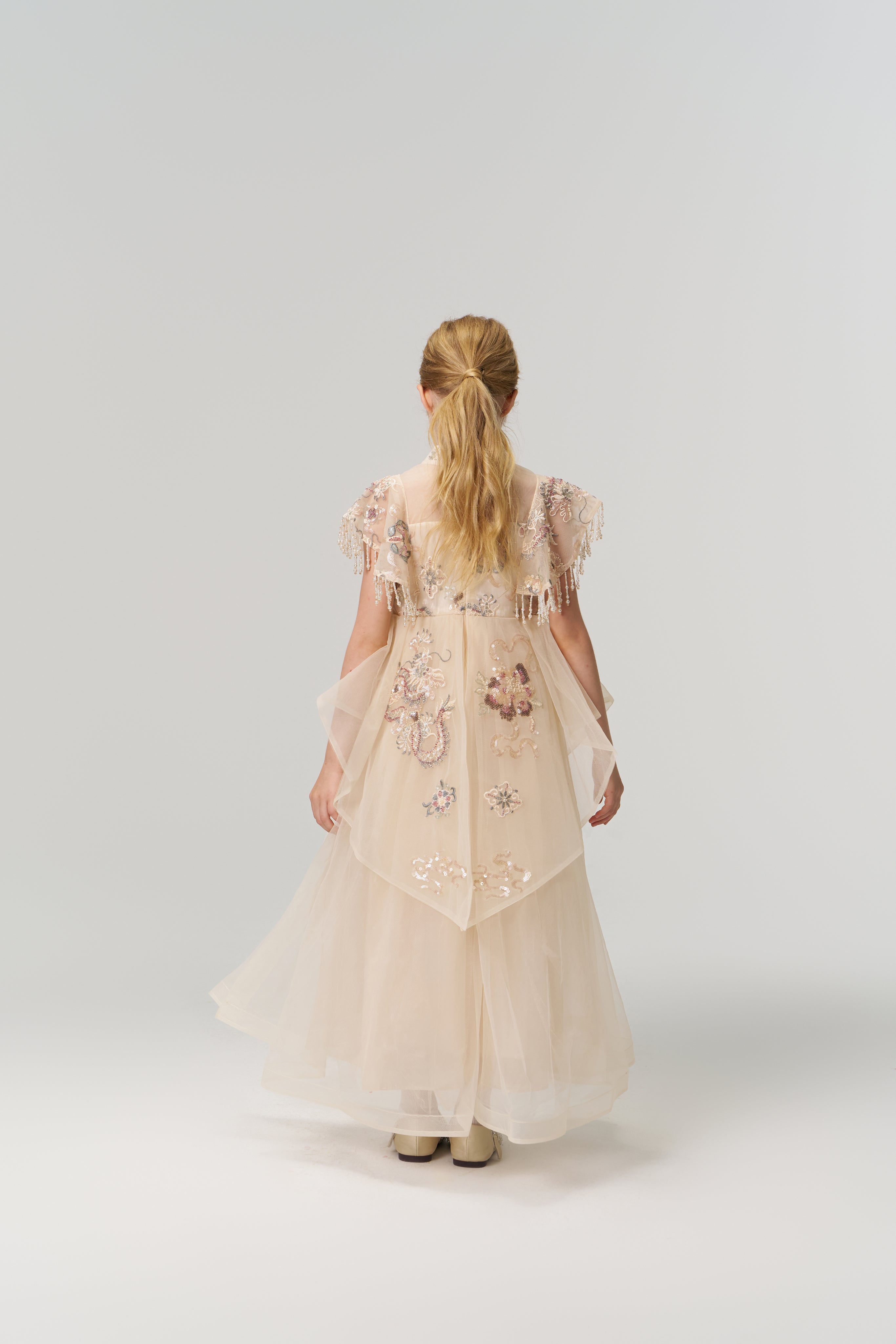 A young girl with blonde hair in a ponytail stands facing away, modeling the KIKI and JOJO Jade Whisper Dress—a long, light pink cheongsam with embroidered florals and a mandarin collar—against a plain white background.