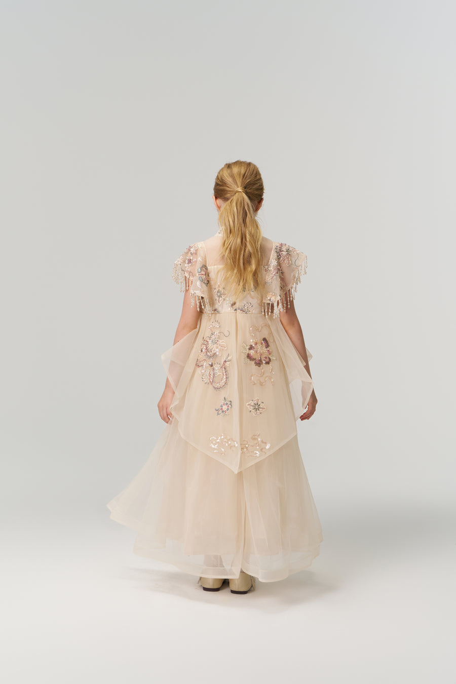 A young girl with blonde hair in a ponytail stands facing away, modeling the KIKI and JOJO Jade Whisper Dress—a long, light pink cheongsam with embroidered florals and a mandarin collar—against a plain white background.