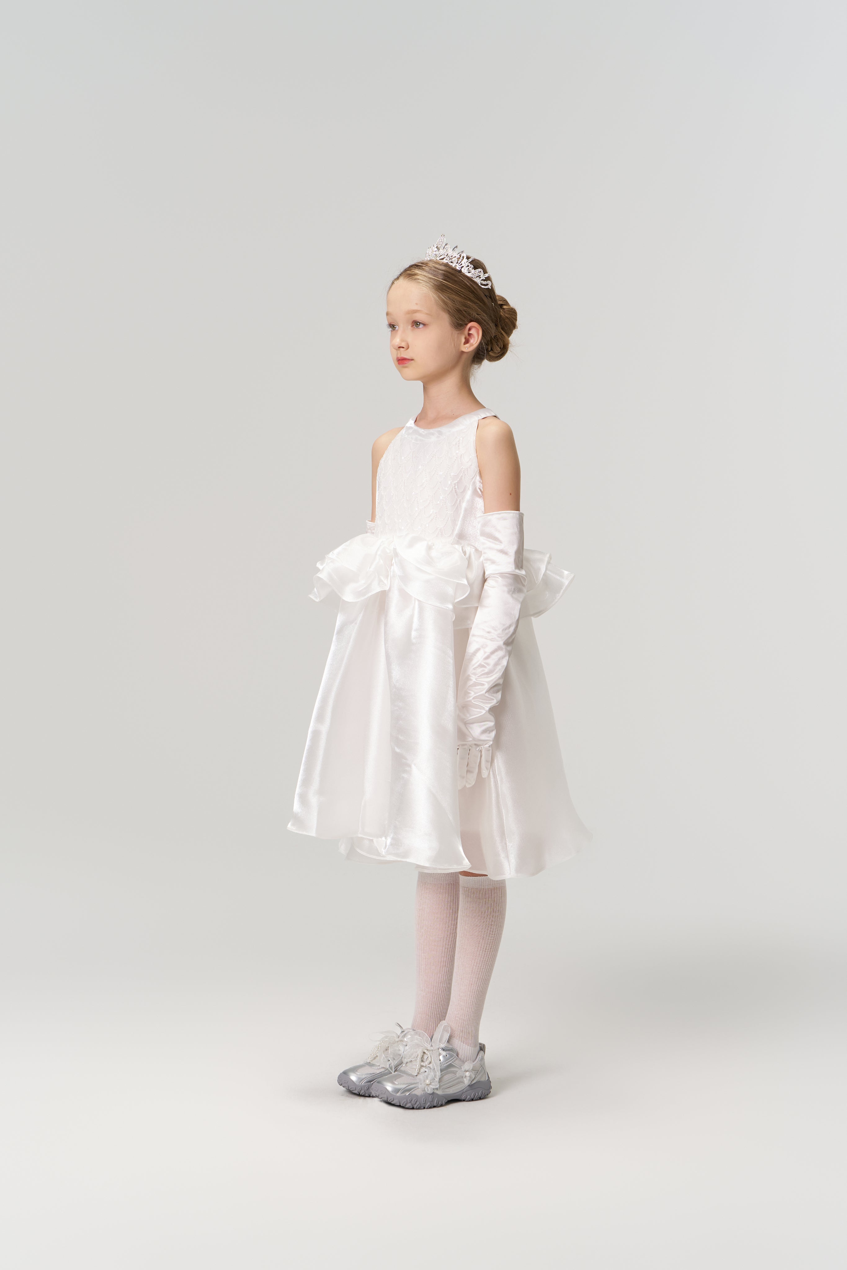 A young girl wears the Pearl Glow Whisper Dress by KIKI and JOJO—a princess-style A-line with beaded embroidery—paired with gloves, a tiara, and silver shoes, standing against a plain light background, facing slightly left.