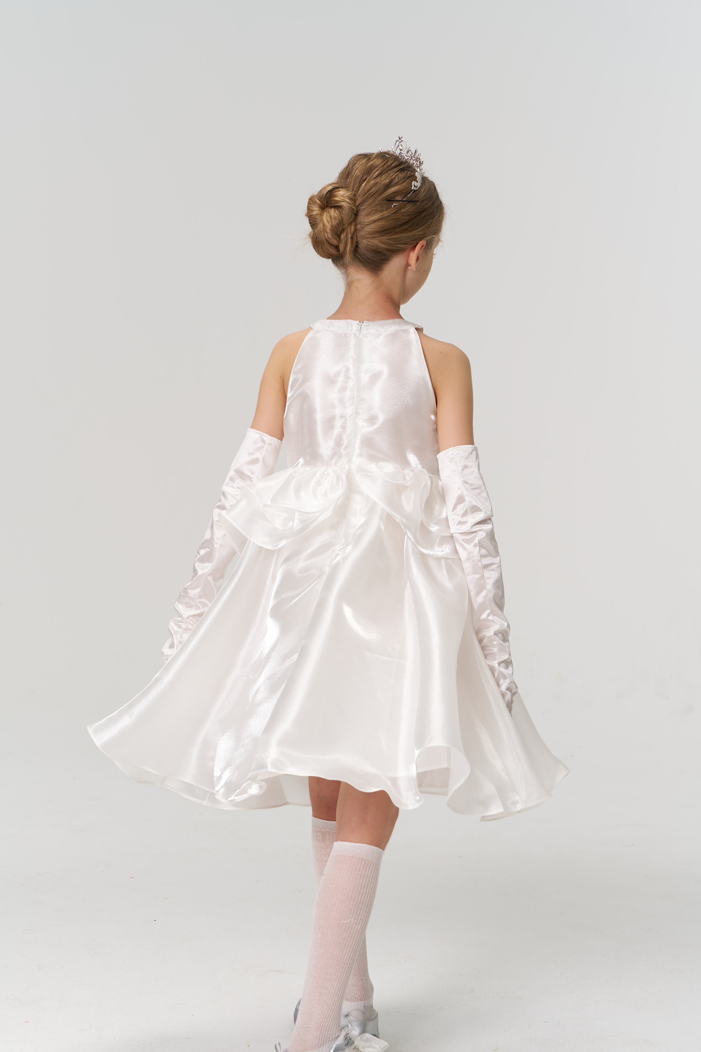A young girl wears the KIKI and JOJO Pearl Glow Whisper Dress, styled a-line with satin fabric, long white gloves, and a tiara, standing with her back to the camera against a simple light background.