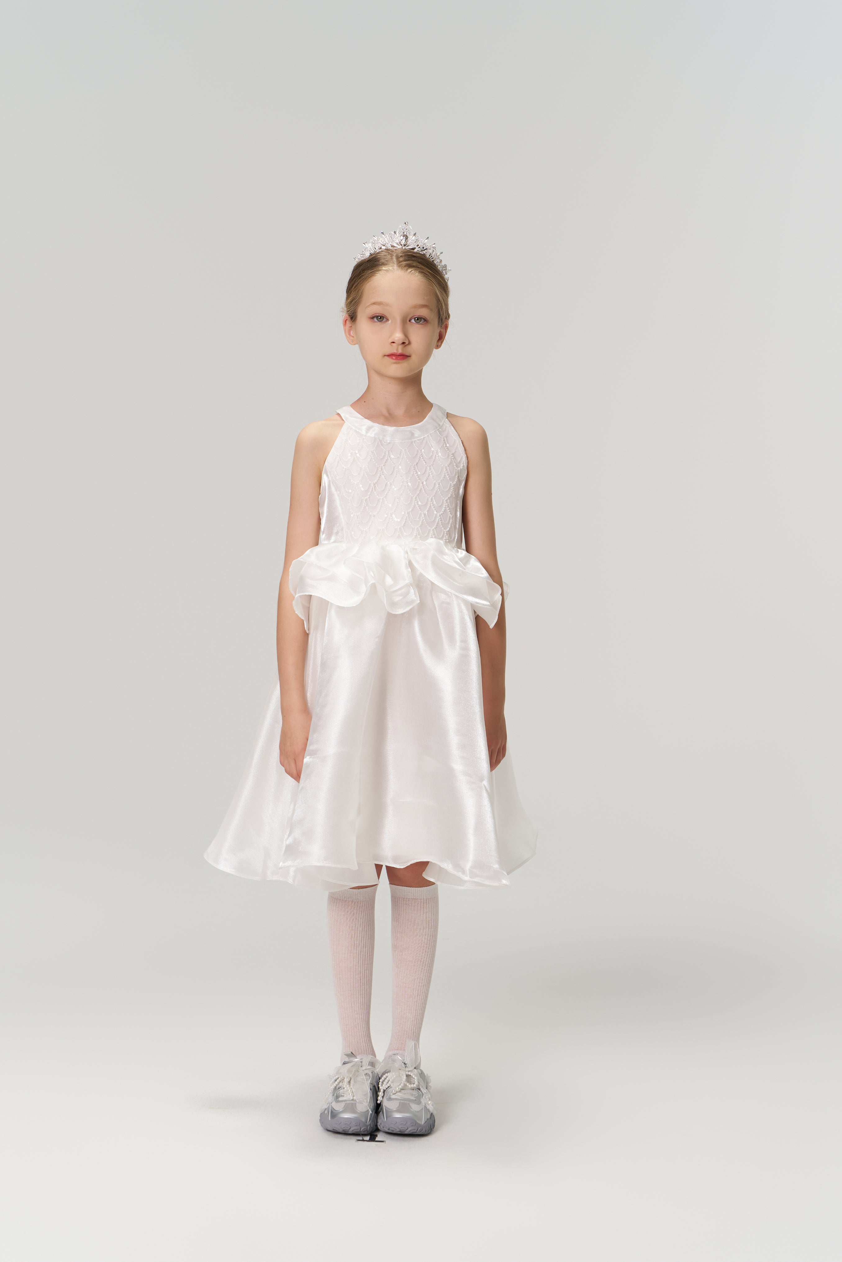 A young girl stands facing forward in the Pearl Glow Whisper Dress by KIKI and JOJO, featuring beaded embroidery. She wears a tiara, knee-high socks, and gray sneakers, set against a plain light background.