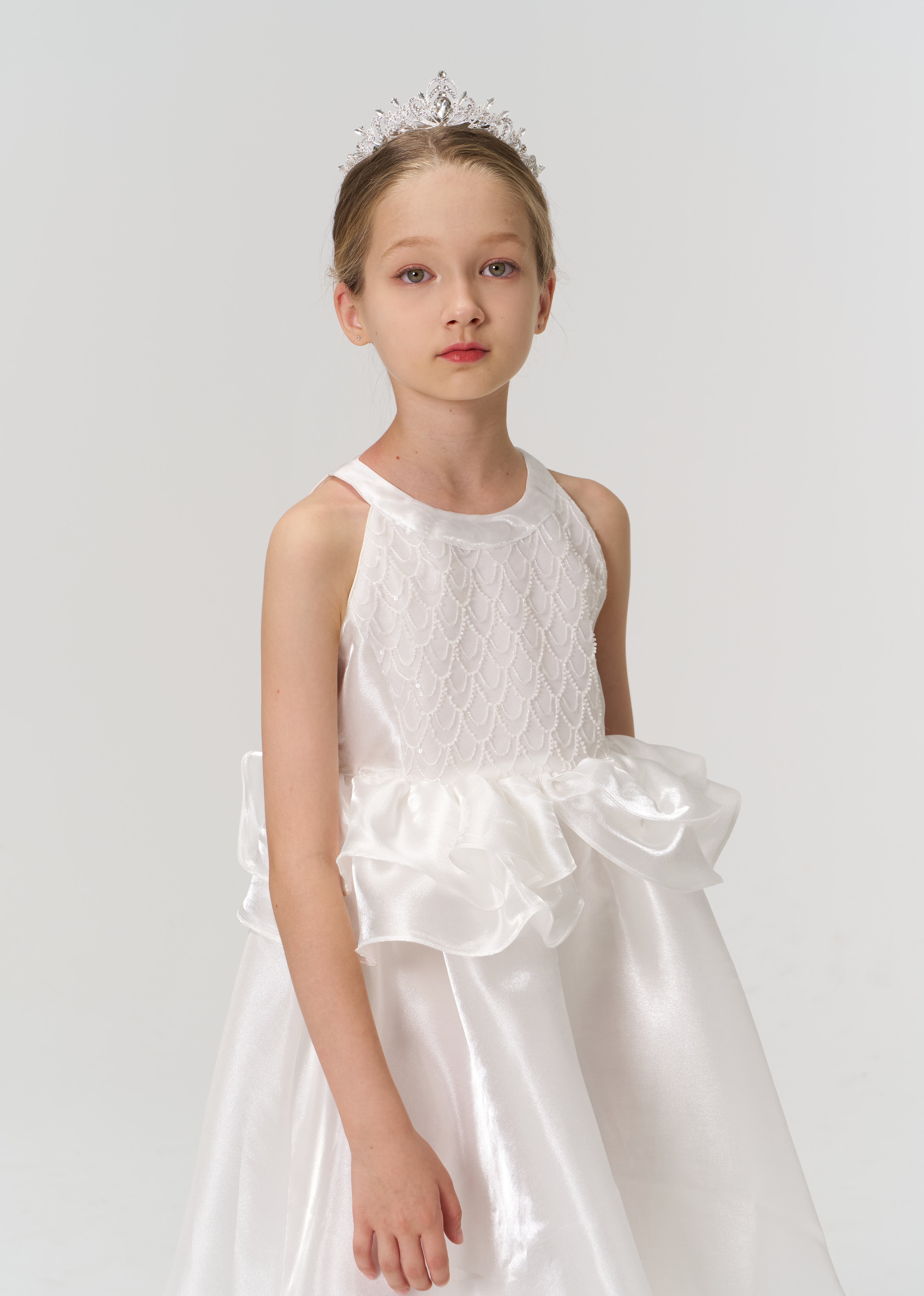 A young girl wearing a white dress with a ruffled waist and a tiara stands against a plain light background, looking directly at the camera.