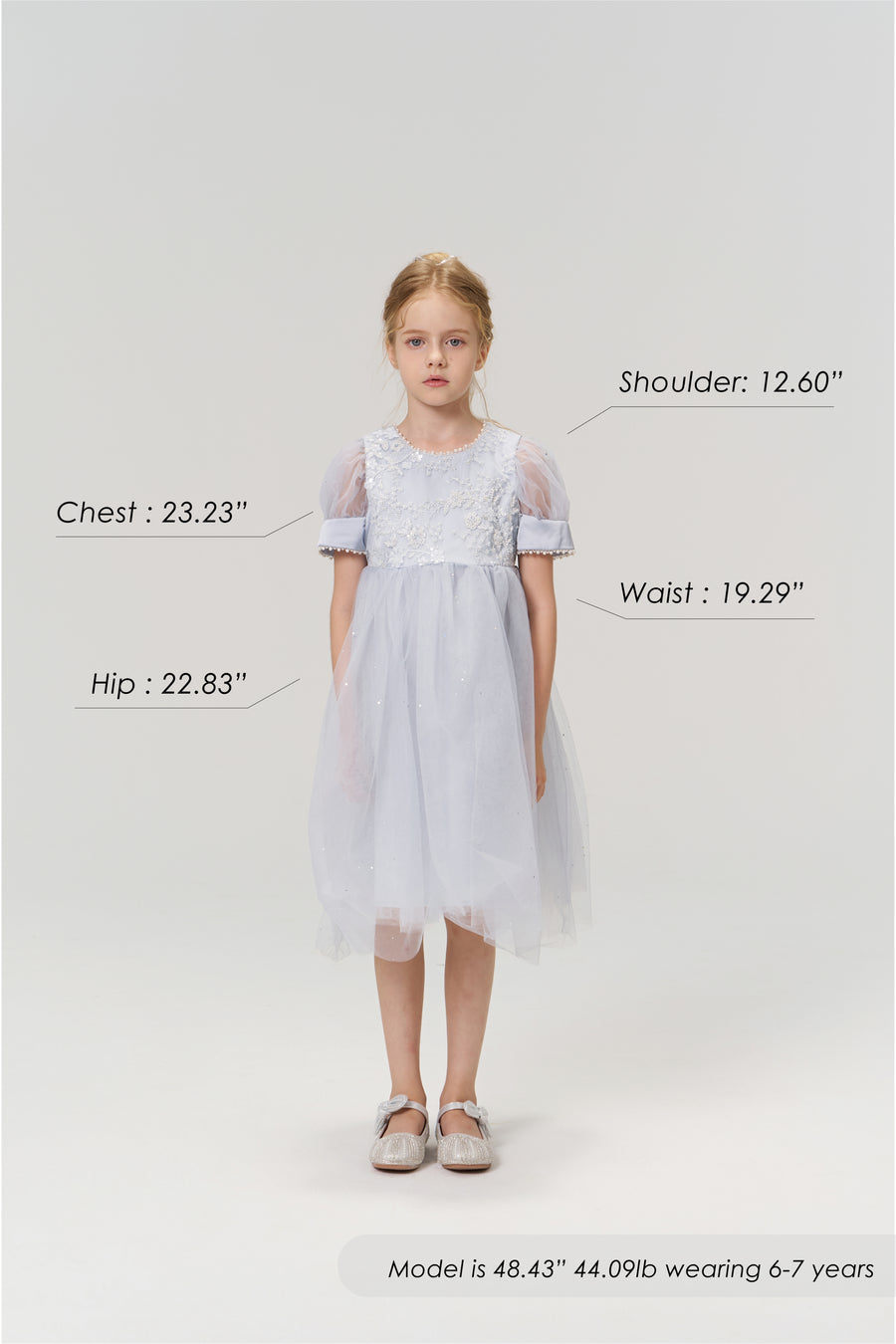 A young girl aged 6-7 models the Mist Whisper Dress by KIKI and JOJO in light blue with silver shoes, standing against a plain background. Her chest, hip, shoulder, and waist measurements are labeled for size reference.