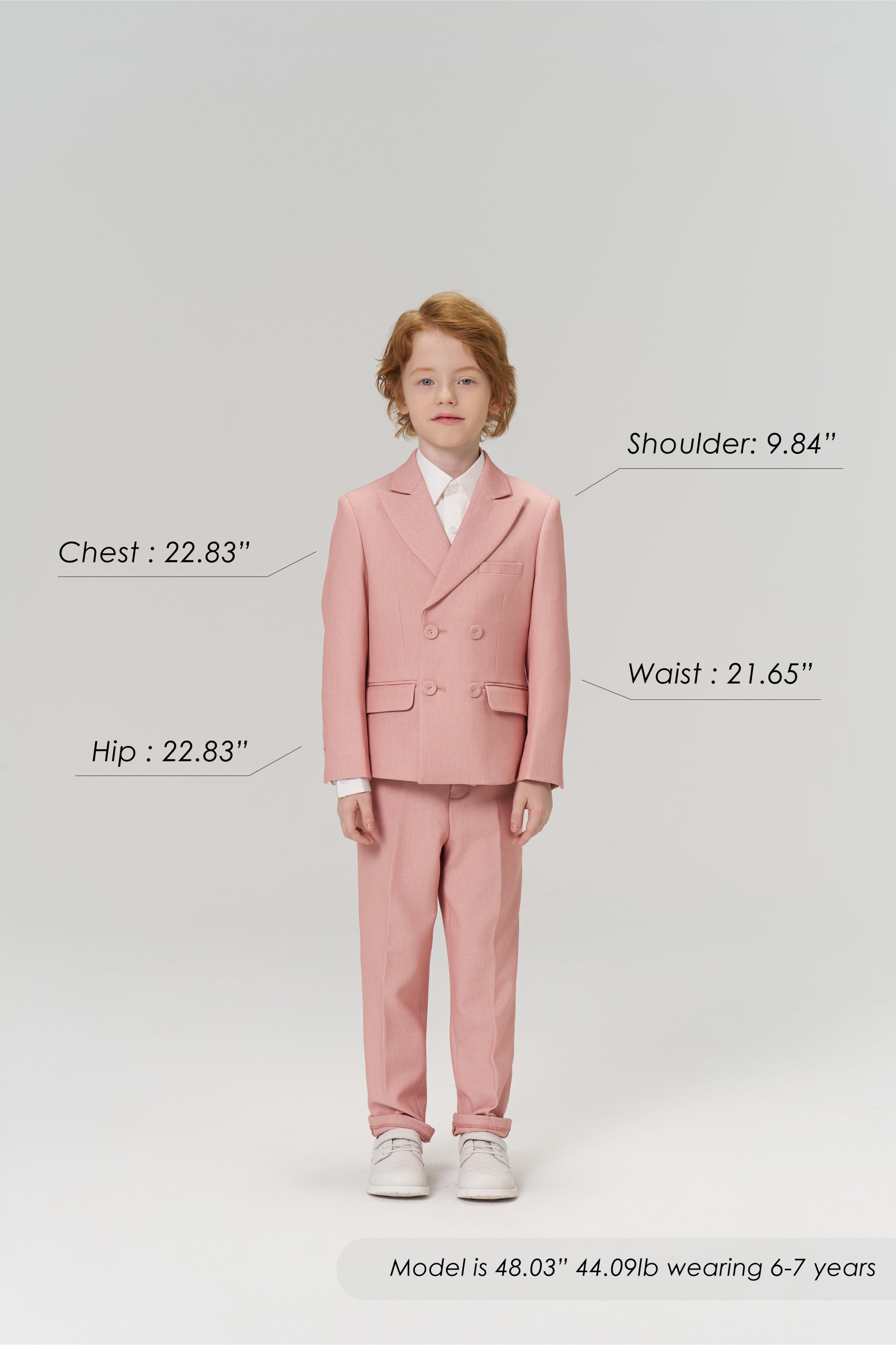 A young boy wears KIKI and JOJO Blush Reverie Suit Pants, posing forward to display the boys’ formal trousers with an adjustable waistband. Model’s size details are shown. Neutral gray background.