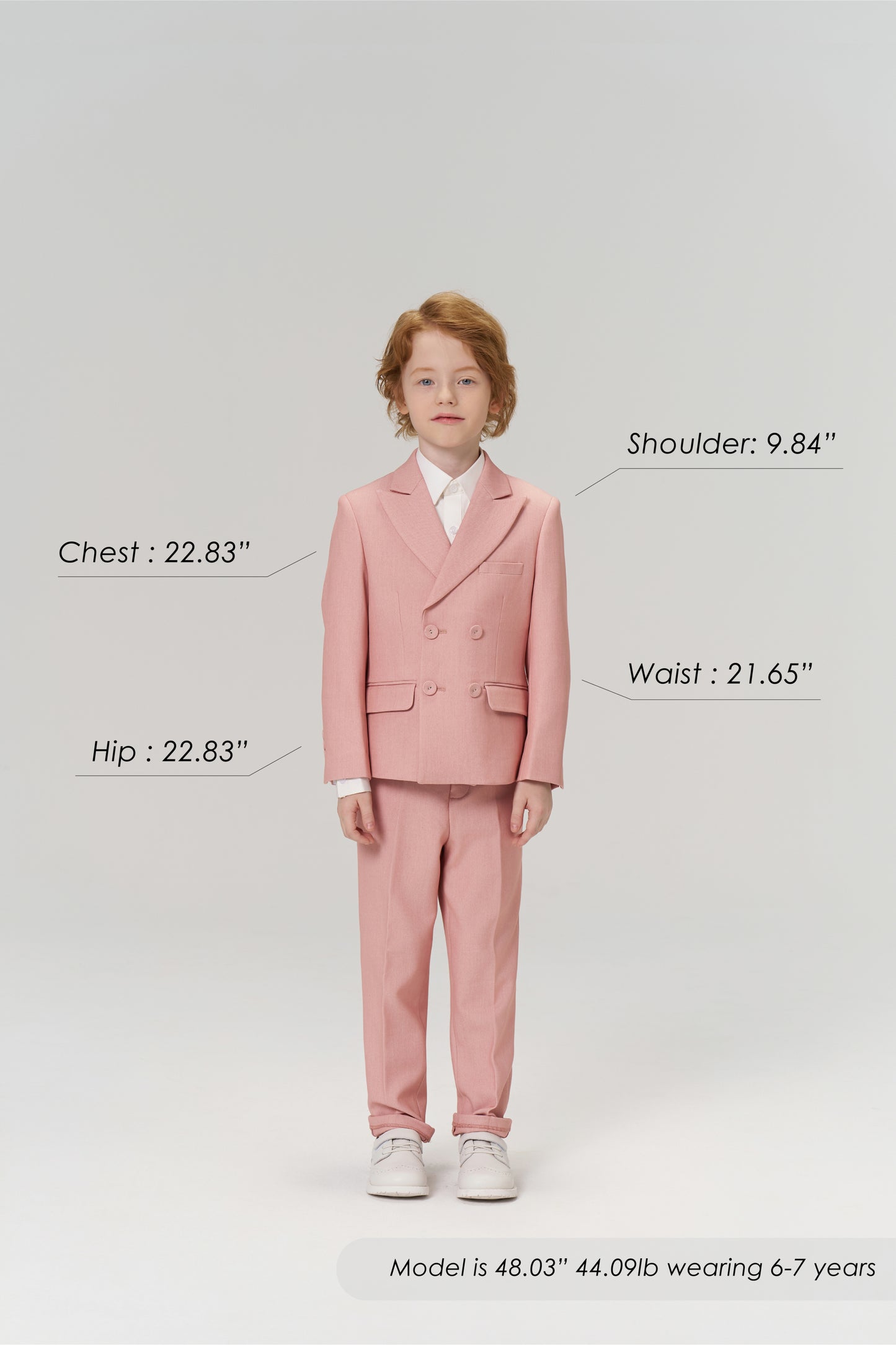 A young boy wears KIKI and JOJO Blush Reverie Suit Pants, posing forward to display the boys’ formal trousers with an adjustable waistband. Model’s size details are shown. Neutral gray background.