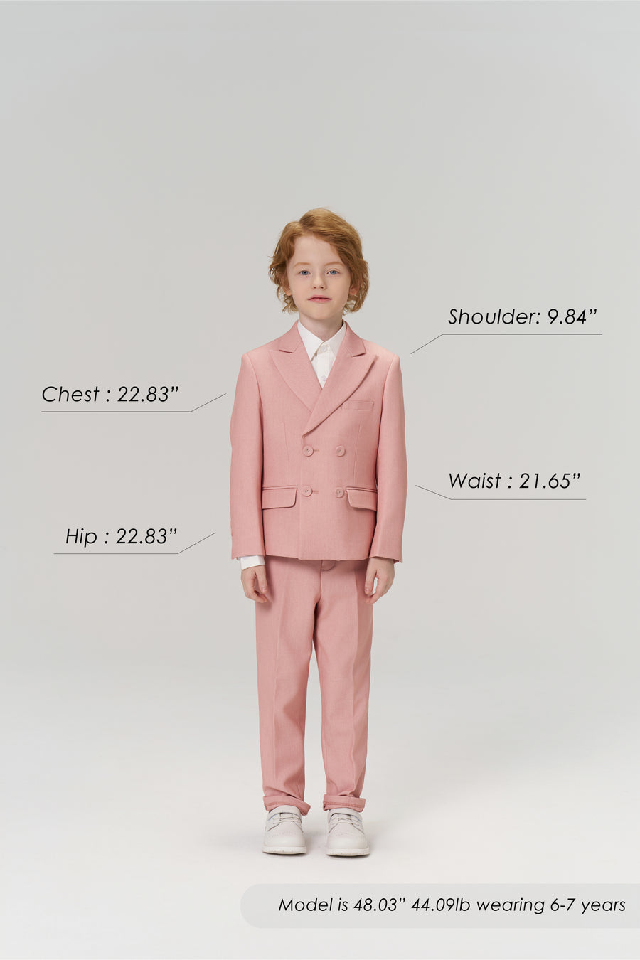 A young boy wears KIKI and JOJO Blush Reverie Suit Pants, posing forward to display the boys’ formal trousers with an adjustable waistband. Model’s size details are shown. Neutral gray background.