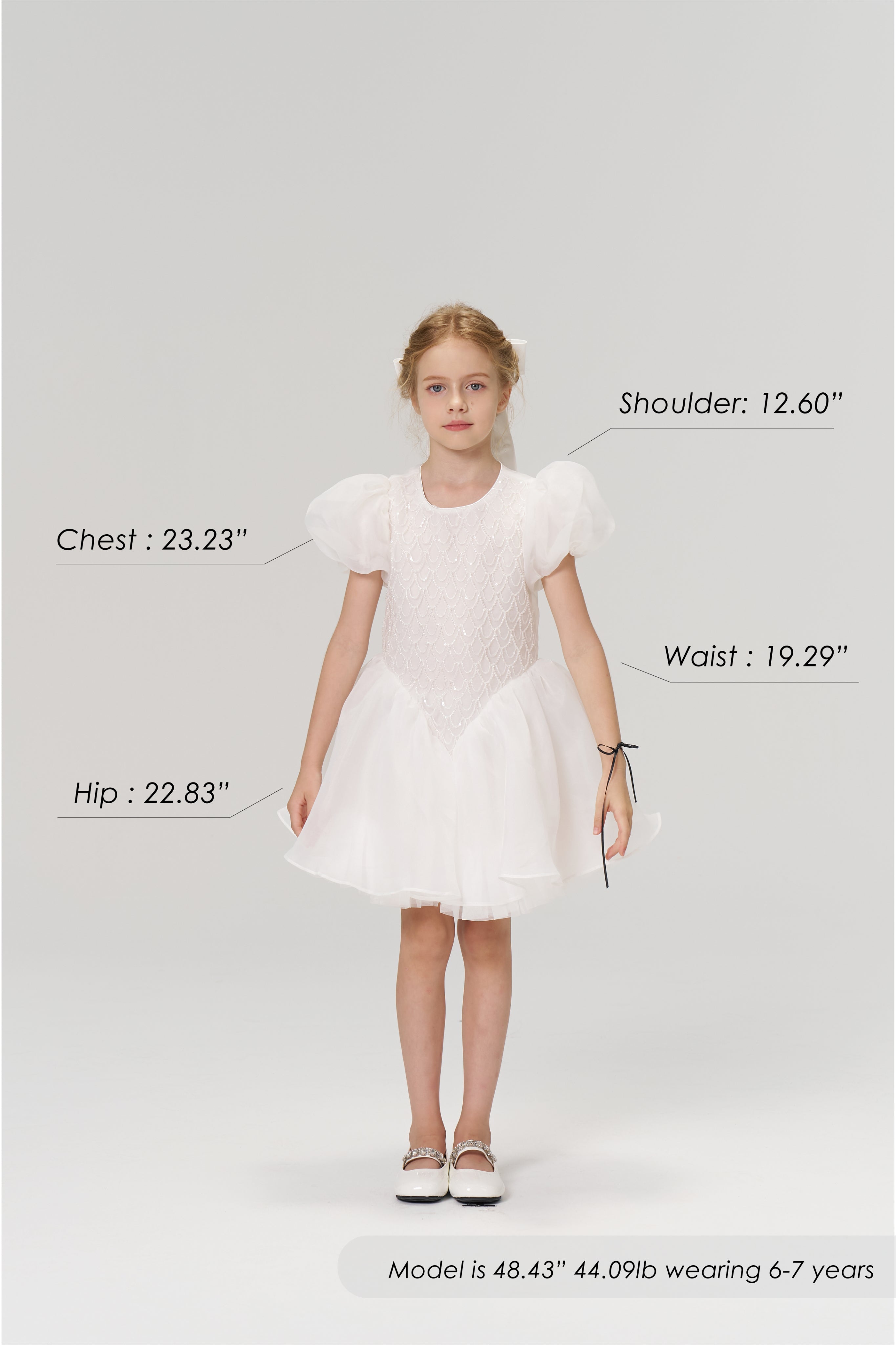 A young girl stands facing forward in the KIKI and JOJO Ballerina Whisper Dress with white shoes; her body measurements, height, weight, and age are displayed around her.