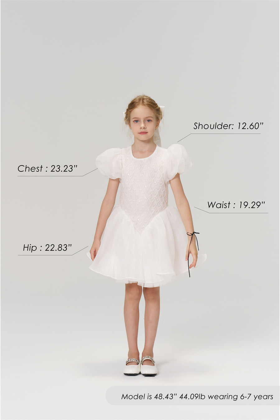A young girl stands facing forward in the KIKI and JOJO Ballerina Whisper Dress with white shoes; her body measurements, height, weight, and age are displayed around her.
