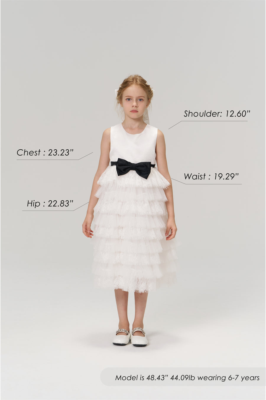 A young girl models the KIKI and JOJO Monochrome Whisper Dress, a black and white layered style with a tulle cake skirt. Shown with size chart for shoulder, chest, waist, and hip. Model is 48.43", 44.09 lb, wearing size 6–7 years.