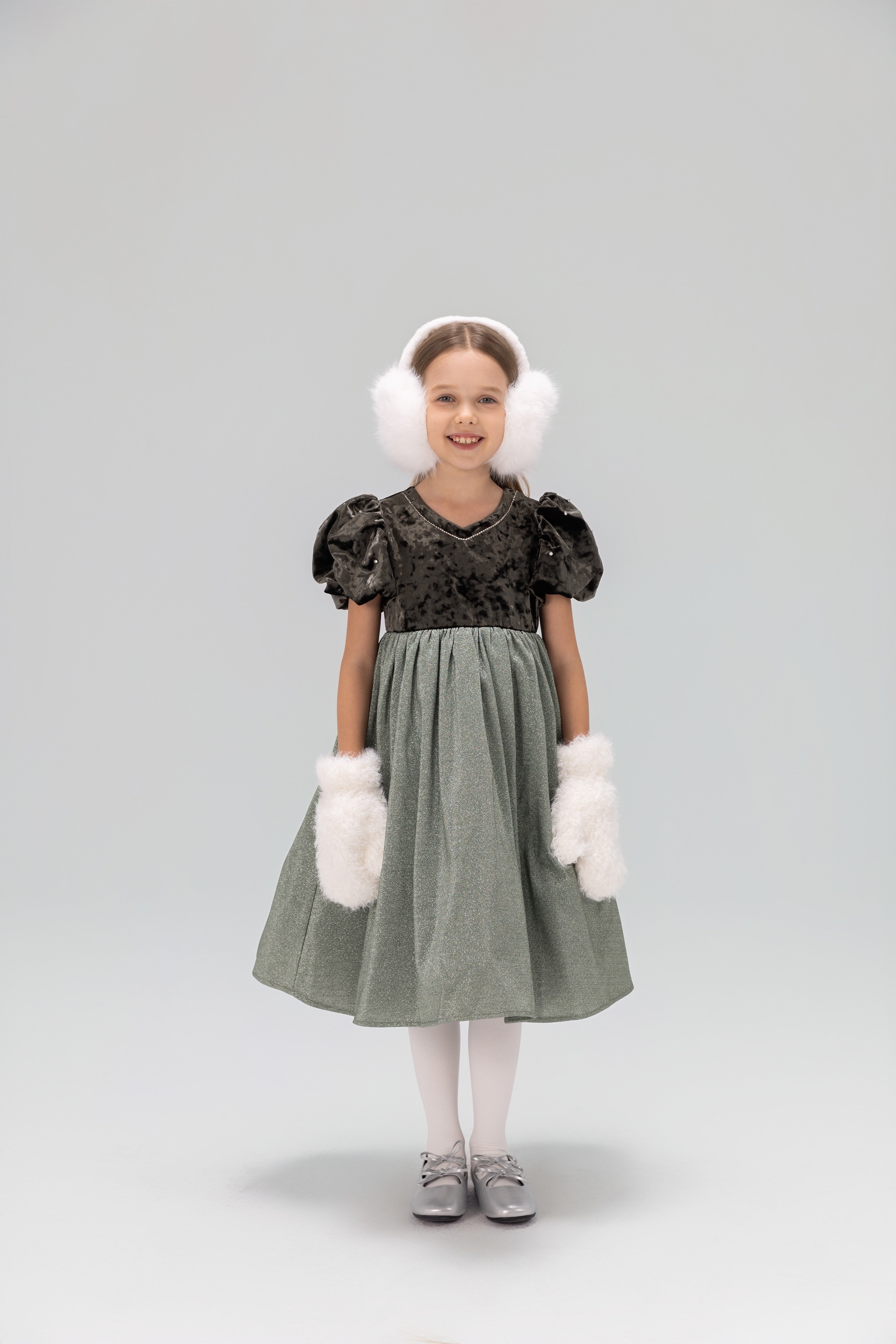 A young girl wears the KIKI and JOJO Girls Velvet Puff Sleeve Dress with Rhinestone Trim in forest green, paired with white earmuffs, fluffy mittens, tights, and gray shoes, smiling at the camera against a plain light background.