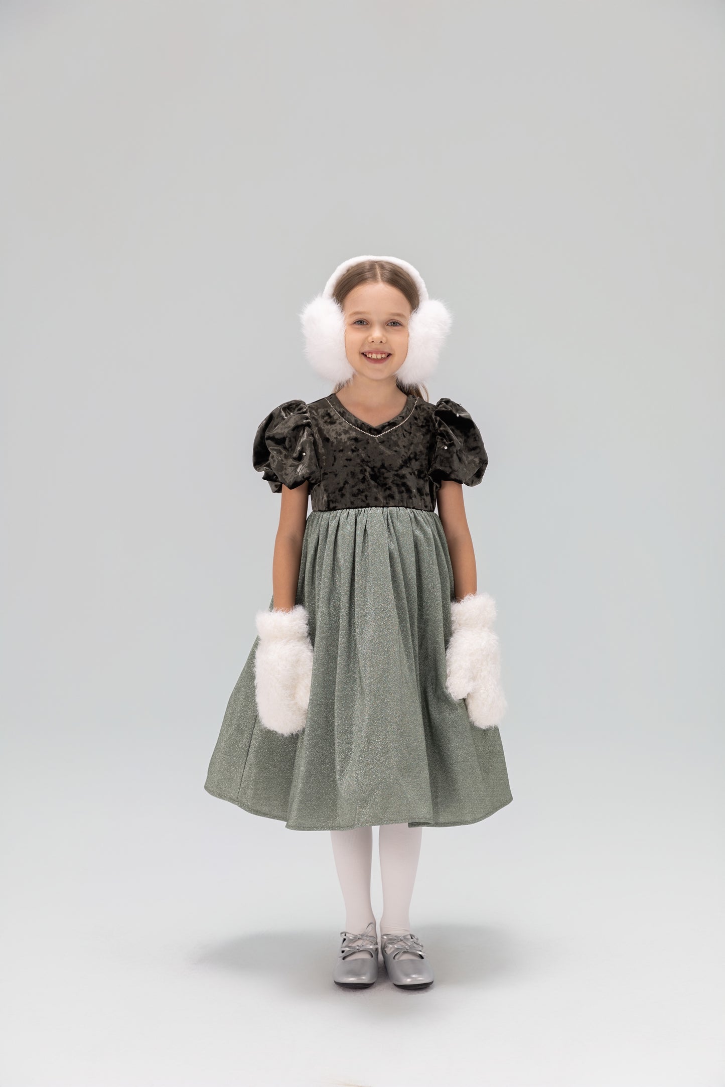 A young girl wears the KIKI and JOJO Girls Velvet Puff Sleeve Dress with Rhinestone Trim in forest green, paired with white earmuffs, fluffy mittens, tights, and gray shoes, smiling at the camera against a plain light background.