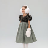 A young girl smiles in a studio, wearing the KIKI and JOJO Girls Velvet Puff Sleeve Dress with Rhinestone Trim in forest green, paired with white tights, silver shoes, white earmuffs, and fluffy mittens, looking slightly to the side.