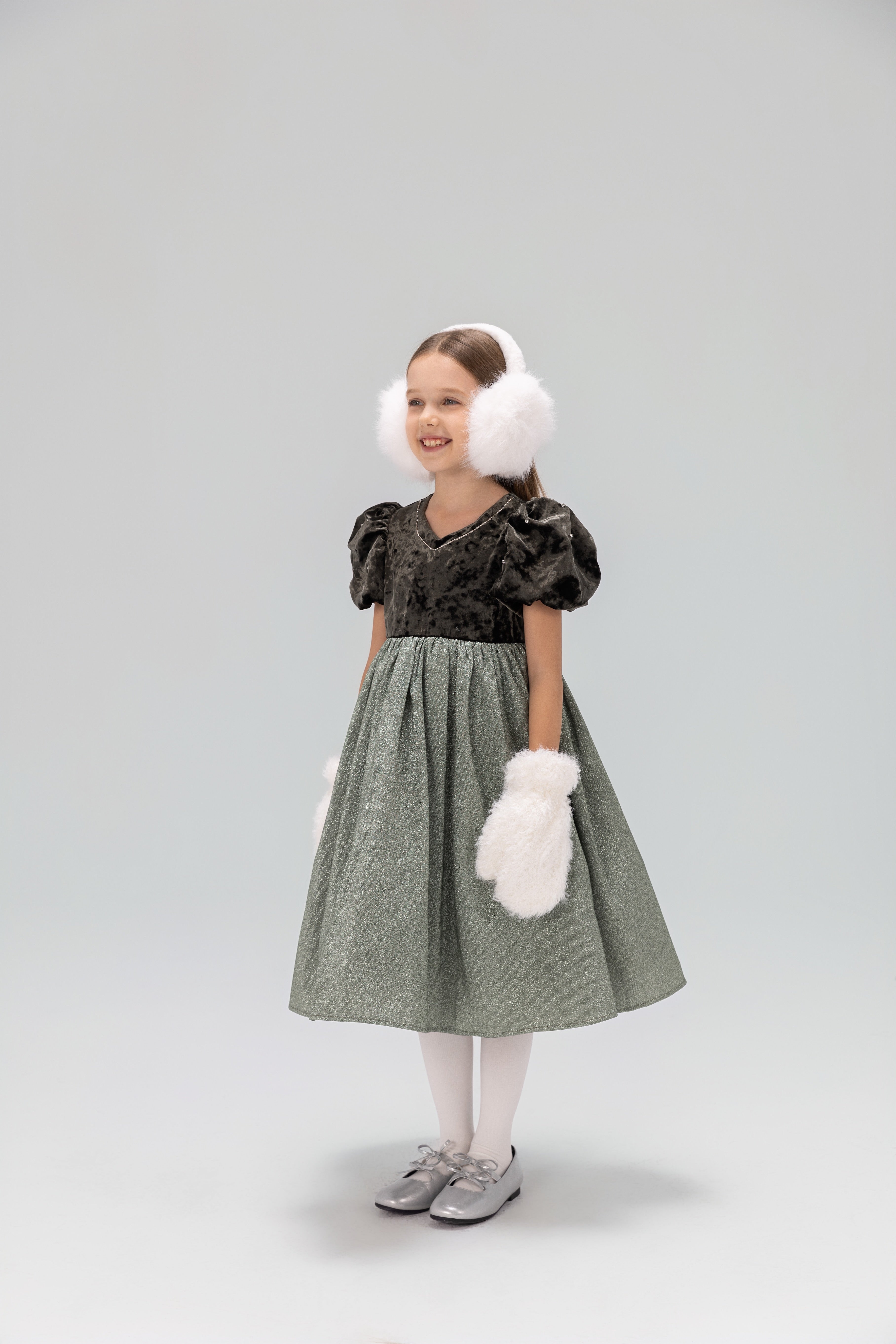 A young girl smiles in a studio, wearing the KIKI and JOJO Girls Velvet Puff Sleeve Dress with Rhinestone Trim in forest green, paired with white tights, silver shoes, white earmuffs, and fluffy mittens, looking slightly to the side.