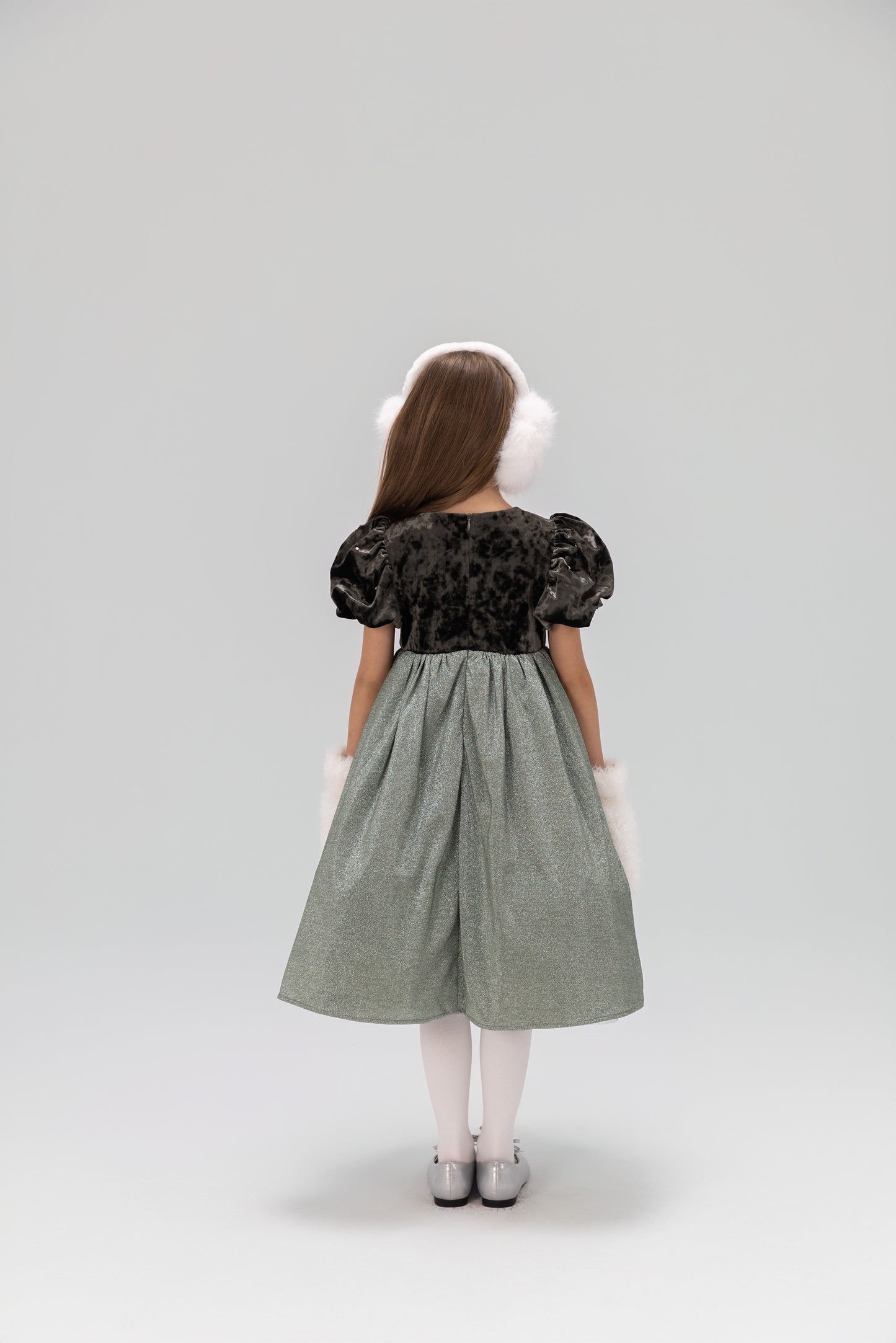 A young girl wears the KIKI and JOJO Girls Velvet Puff Sleeve Dress with Rhinestone Trim in Forest Green, styled with white tights, silver shoes, and white earmuffs and cuffs, standing against a plain background.