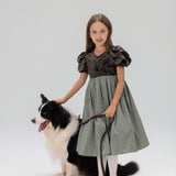 A young girl in a KIKI and JOJO Girls Velvet Puff Sleeve Dress with Rhinestone Trim in forest green stands holding a leash beside a black and white dog, both posed against a plain light background.
