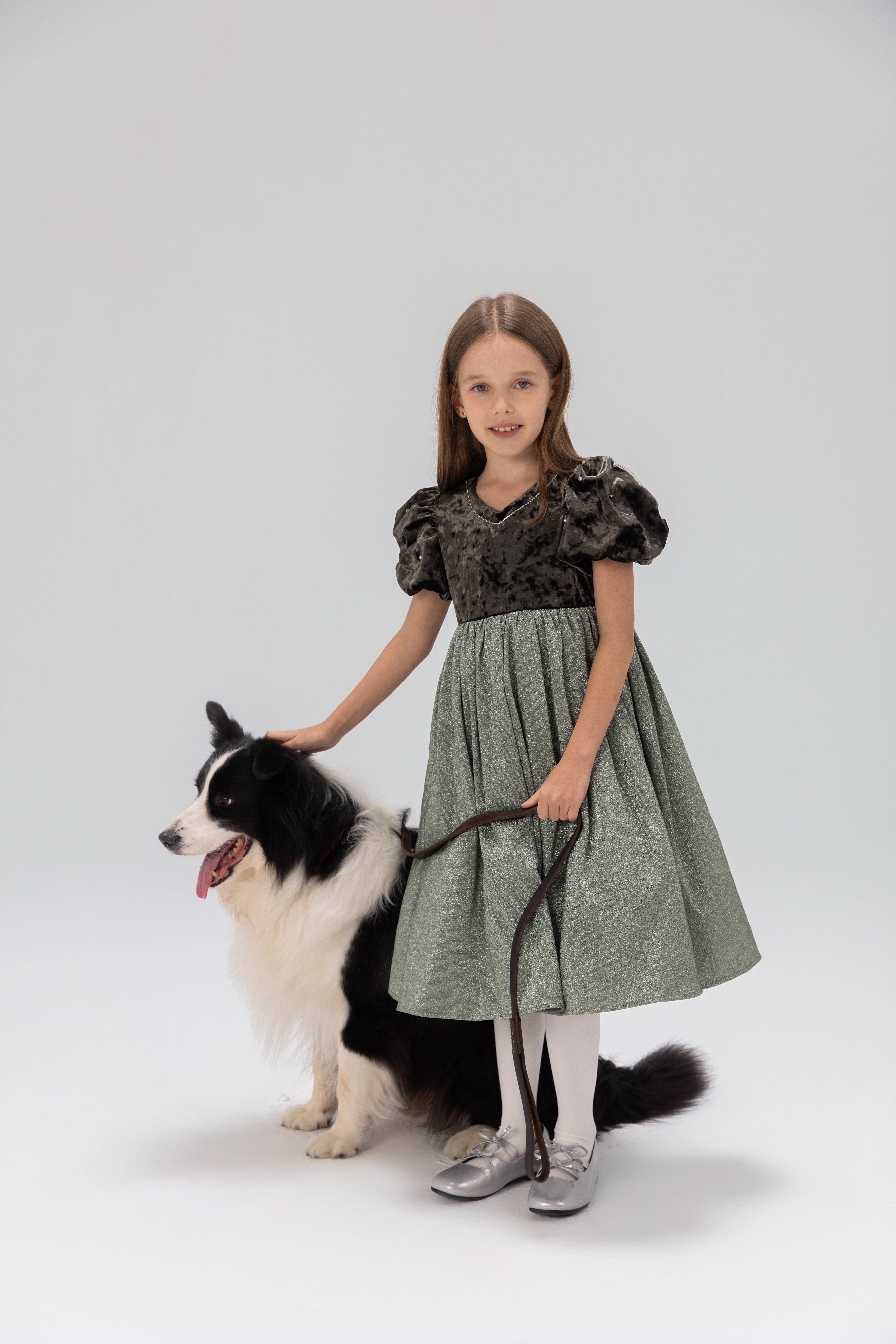 A young girl in a KIKI and JOJO Girls Velvet Puff Sleeve Dress with Rhinestone Trim in forest green stands holding a leash beside a black and white dog, both posed against a plain light background.