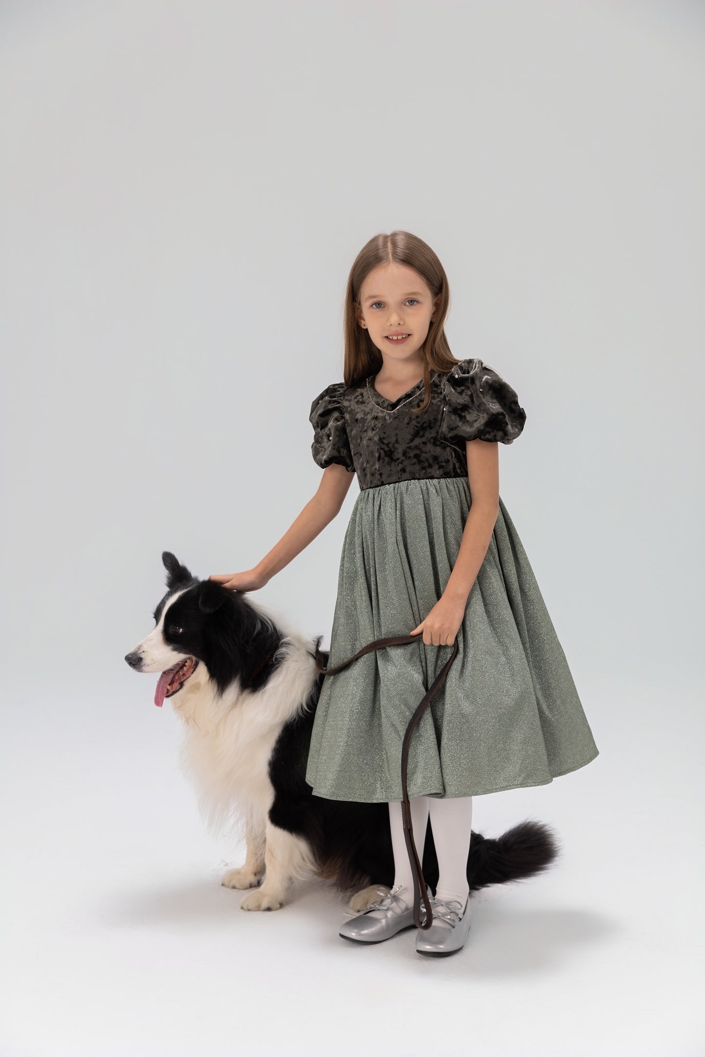 A young girl in a KIKI and JOJO Girls Velvet Puff Sleeve Dress with Rhinestone Trim in forest green stands holding a leash beside a black and white dog, both posed against a plain light background.