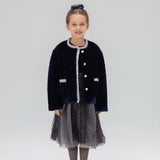 A young girl faces forward in a KIKI and JOJO Girls Quilted Ruffle Jacket in sapphire blue, paired with a gray tulle skirt, gray tights, black shoes, and a matching hair bow against a plain light background.