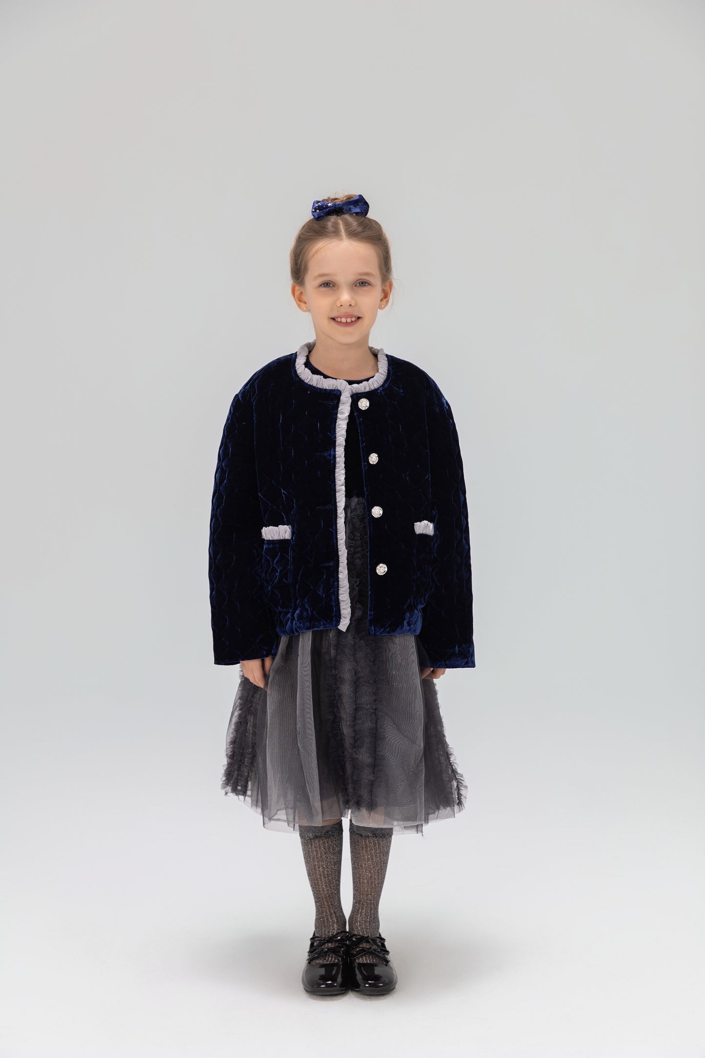 A young girl faces forward in a KIKI and JOJO Girls Quilted Ruffle Jacket in sapphire blue, paired with a gray tulle skirt, gray tights, black shoes, and a matching hair bow against a plain light background.