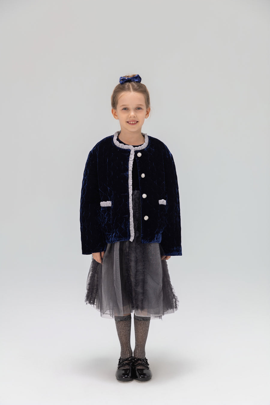 A young girl faces forward in a KIKI and JOJO Girls Quilted Ruffle Jacket in sapphire blue, paired with a gray tulle skirt, gray tights, black shoes, and a matching hair bow against a plain light background.