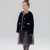 A young girl smiles in a KIKI and JOJO Girls Quilted Ruffle Jacket in Sapphire Blue, paired with a tulle skirt, patterned tights, black shoes, and a matching blue hair accessory against a plain light background.
