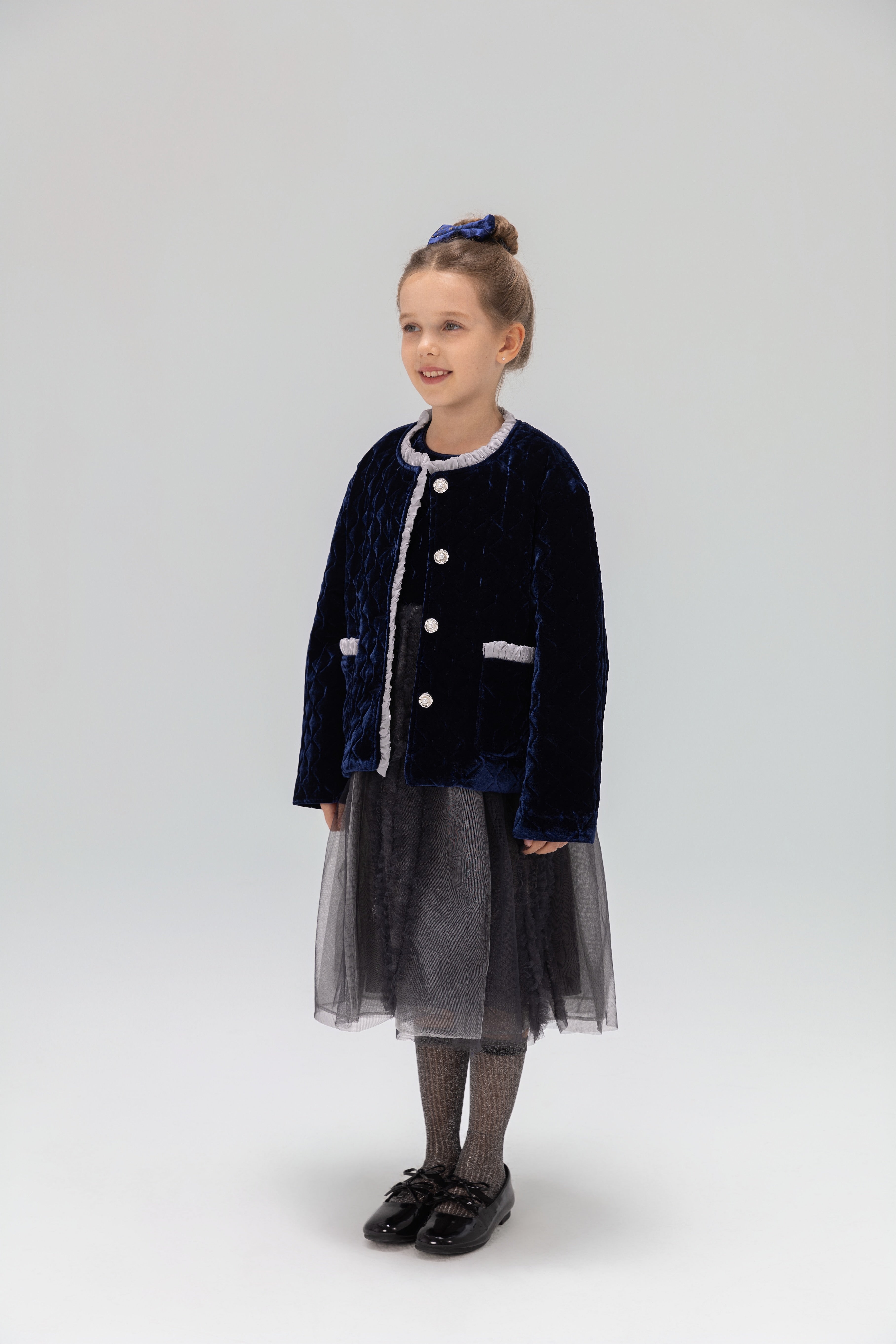 A young girl smiles in a KIKI and JOJO Girls Quilted Ruffle Jacket in Sapphire Blue, paired with a tulle skirt, patterned tights, black shoes, and a matching blue hair accessory against a plain light background.