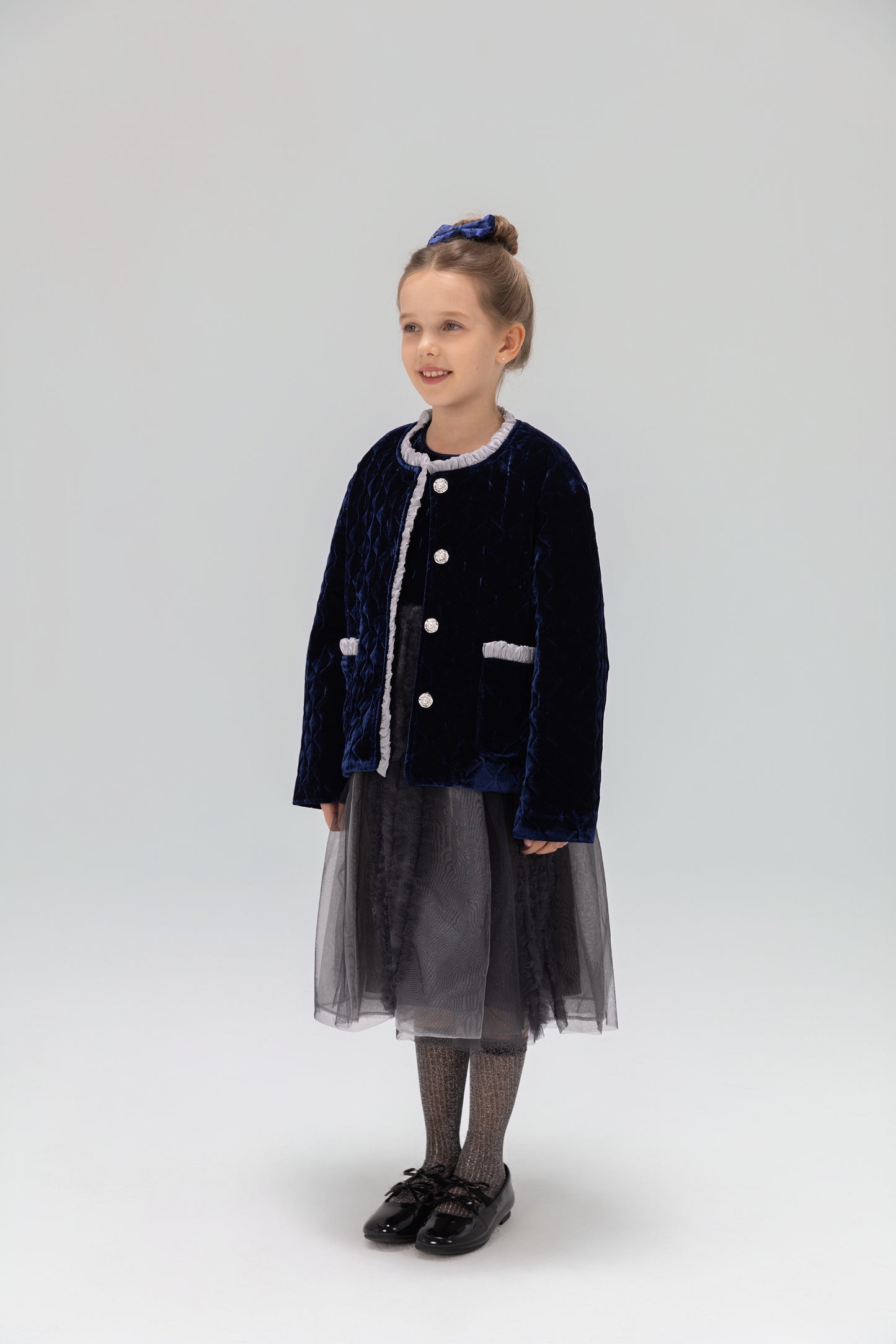 A young girl smiles in a KIKI and JOJO Girls Quilted Ruffle Jacket in Sapphire Blue, paired with a tulle skirt, patterned tights, black shoes, and a matching blue hair accessory against a plain light background.