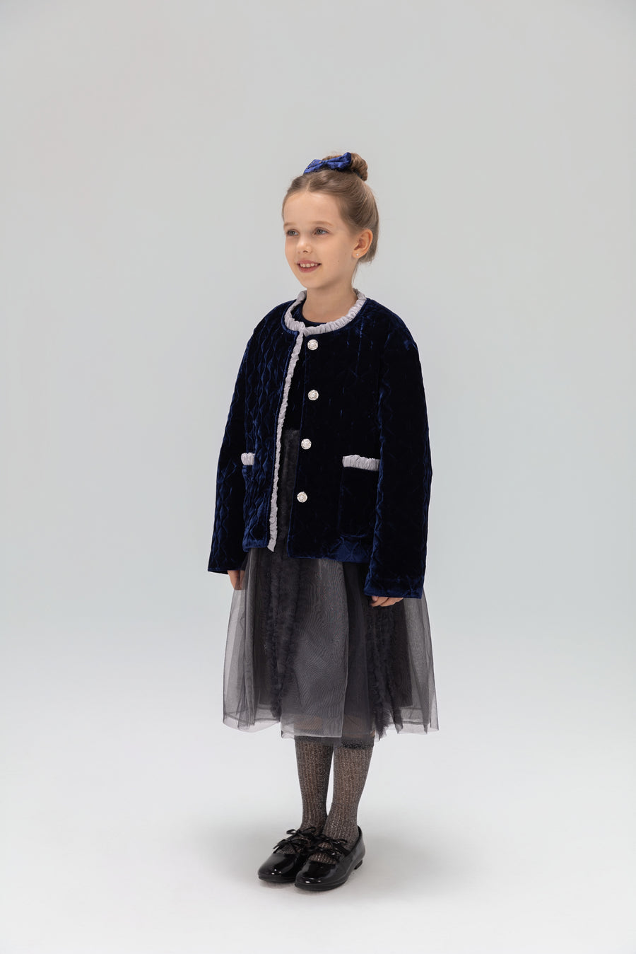 A young girl smiles in a KIKI and JOJO Girls Quilted Ruffle Jacket in Sapphire Blue, paired with a tulle skirt, patterned tights, black shoes, and a matching blue hair accessory against a plain light background.