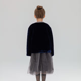 A young girl, seen from behind, wears the KIKI and JOJO Girls Quilted Ruffle Jacket in sapphire blue with a gray tulle skirt, patterned tights, and black shoes against a light backdrop.