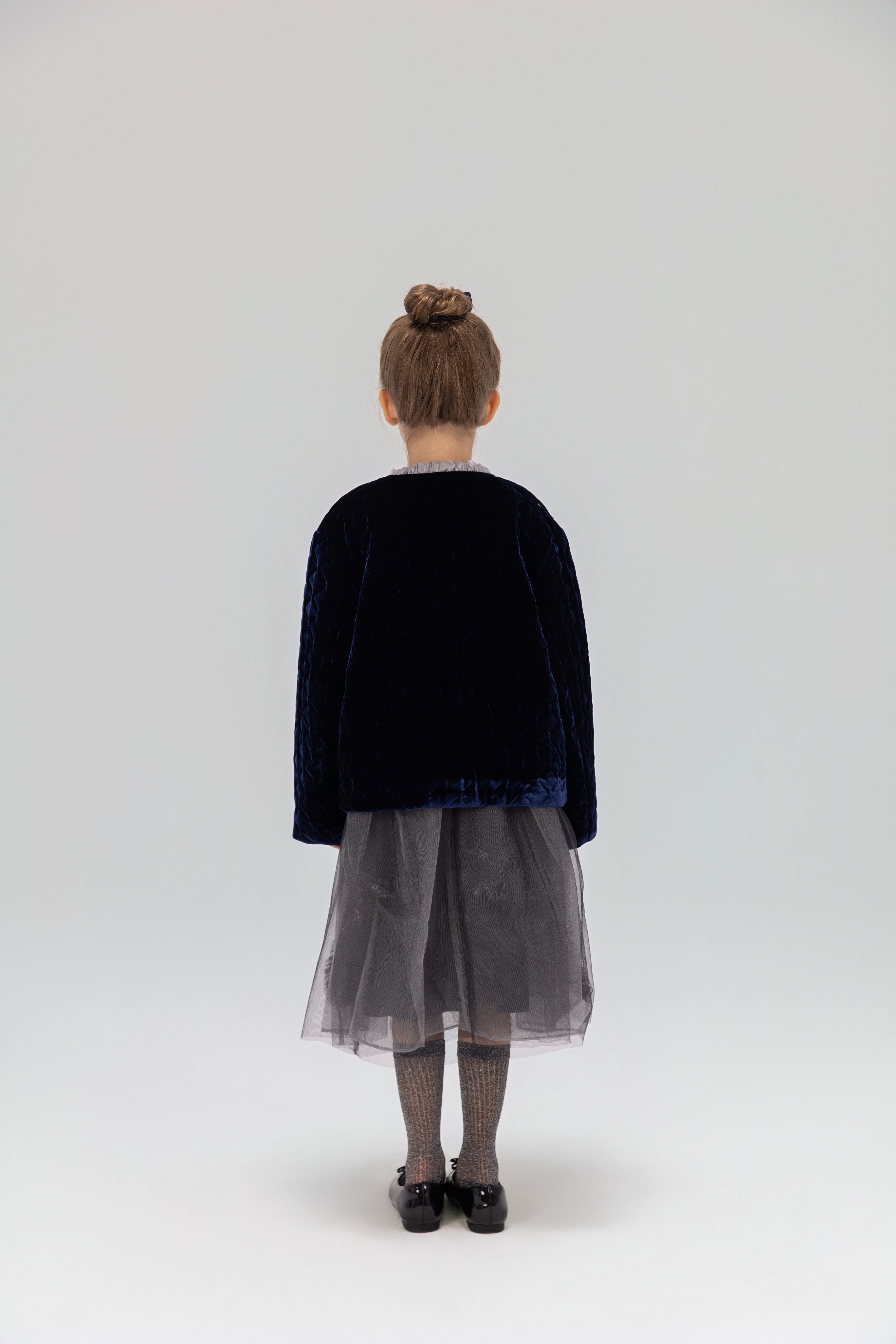 A young girl, seen from behind, wears the KIKI and JOJO Girls Quilted Ruffle Jacket in sapphire blue with a gray tulle skirt, patterned tights, and black shoes against a light backdrop.
