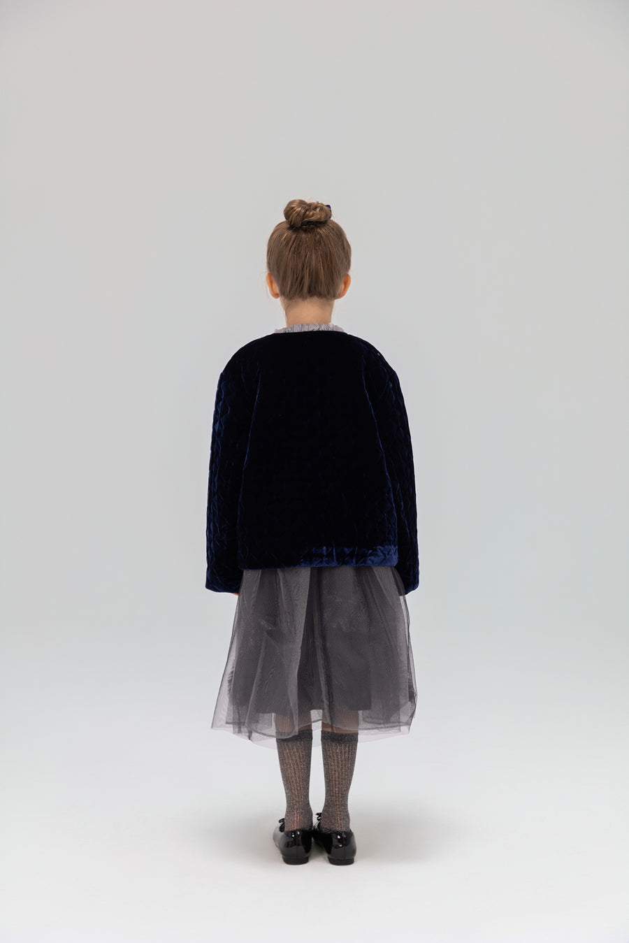 A young girl, seen from behind, wears the KIKI and JOJO Girls Quilted Ruffle Jacket in sapphire blue with a gray tulle skirt, patterned tights, and black shoes against a light backdrop.