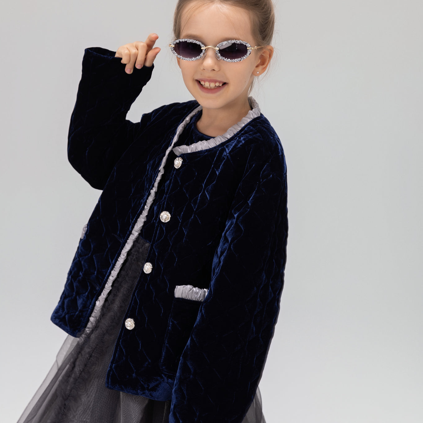 A young girl models the KIKI and JOJO Girls Quilted Ruffle Jacket in sapphire blue, paired with a tulle dress, sunglasses, and a matching hair bow against a plain light background.
