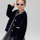 A young girl models the KIKI and JOJO Girls Quilted Ruffle Jacket in sapphire blue, paired with a tulle dress, sunglasses, and a matching hair bow against a plain light background.