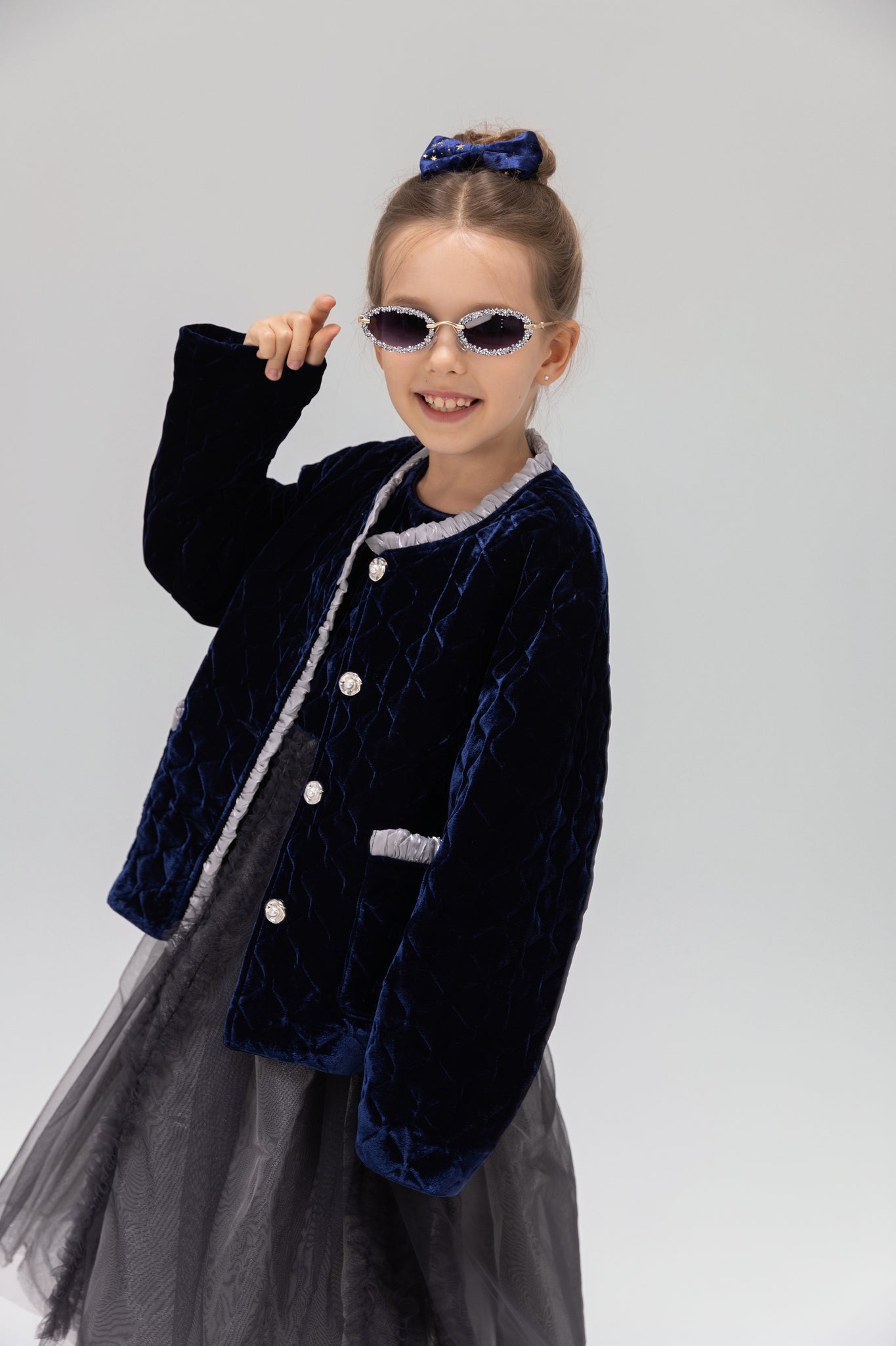 A young girl models the KIKI and JOJO Girls Quilted Ruffle Jacket in sapphire blue, paired with a tulle dress, sunglasses, and a matching hair bow against a plain light background.