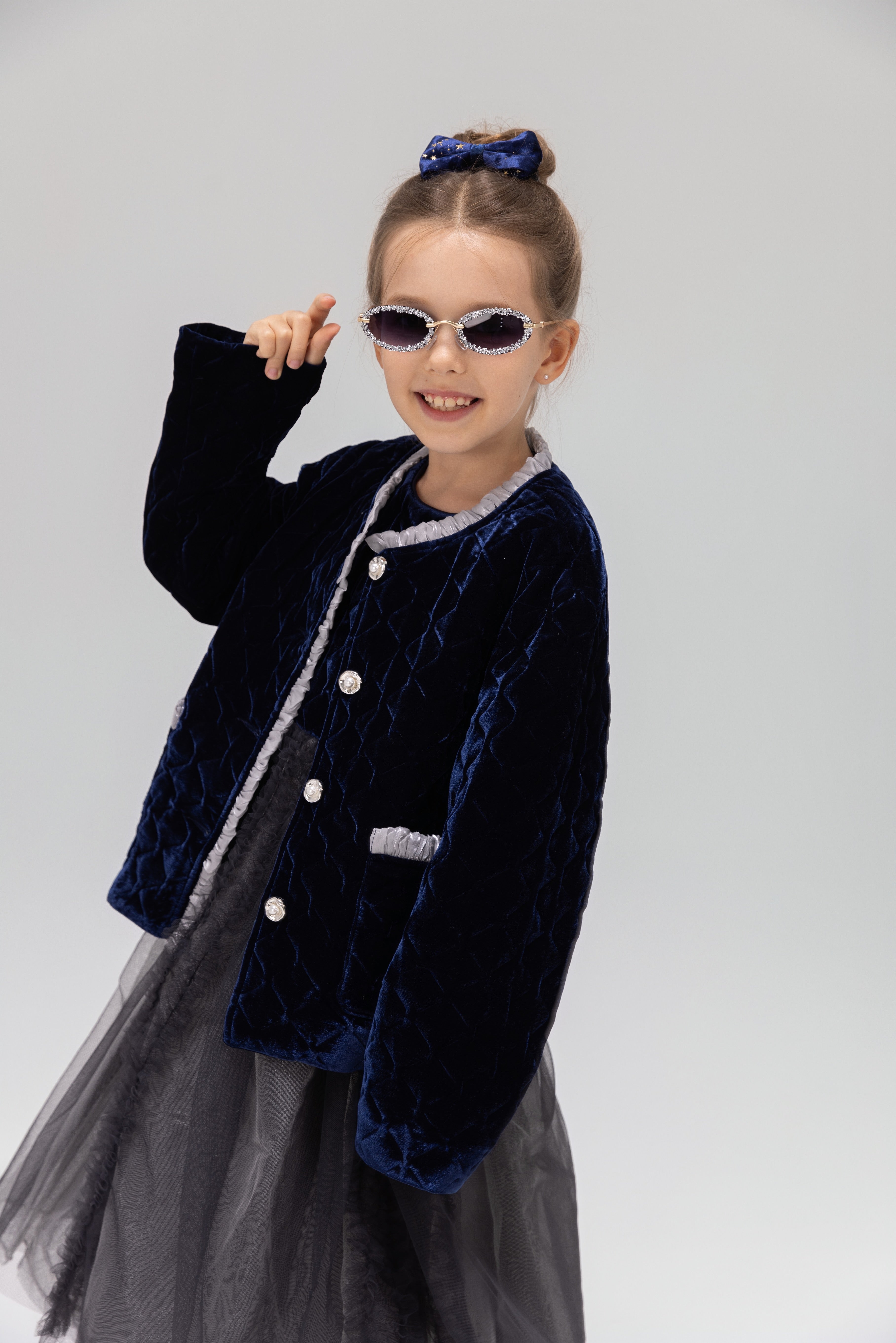 A young girl models the KIKI and JOJO Girls Quilted Ruffle Jacket in sapphire blue, paired with a tulle dress, sunglasses, and a matching hair bow against a plain light background.