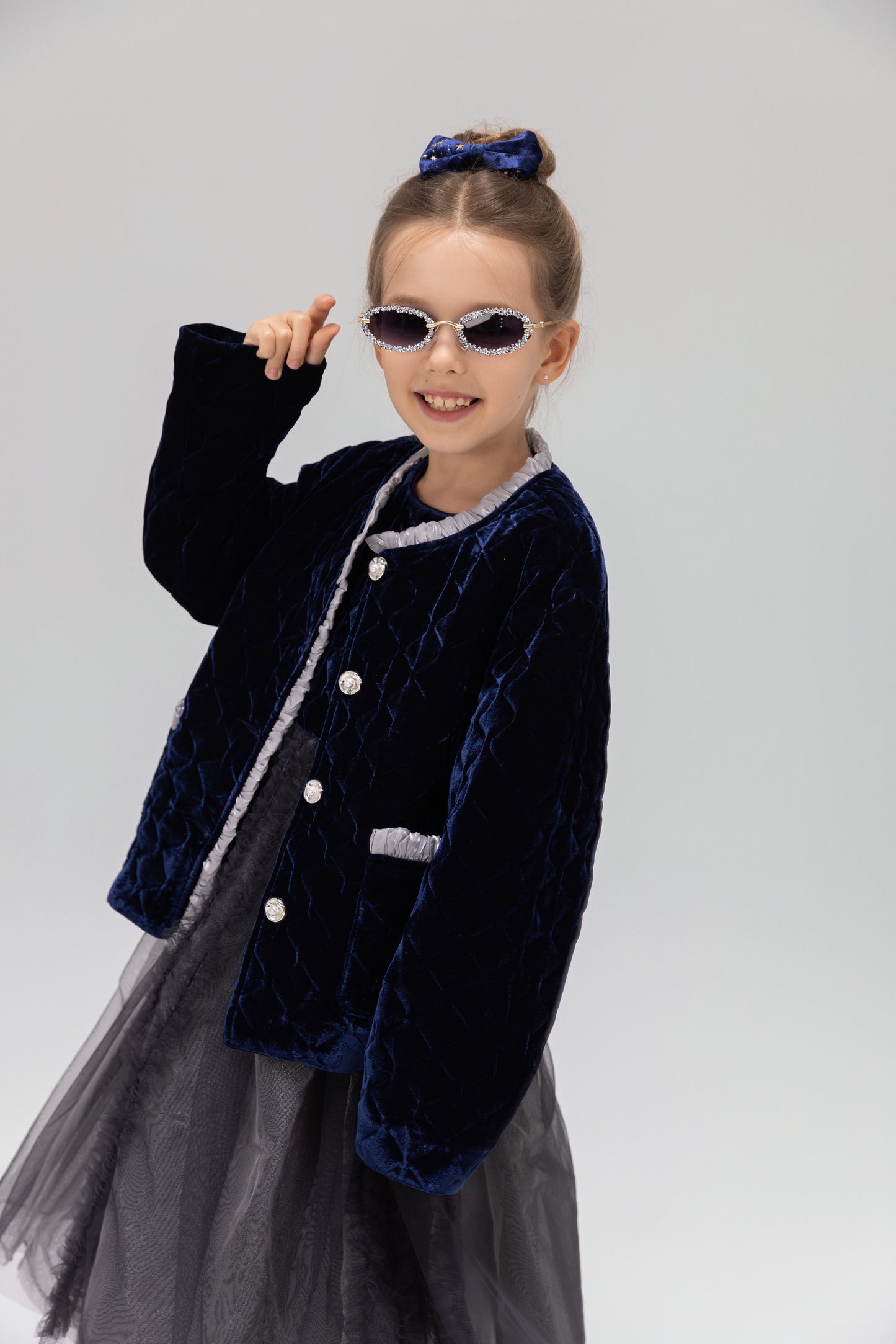 A young girl models the KIKI and JOJO Girls Quilted Ruffle Jacket in sapphire blue, paired with a tulle dress, sunglasses, and a matching hair bow against a plain light background.