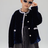 A young girl wearing sunglasses, a gray dress, and the KIKI and JOJO Girls Quilted Ruffle Jacket in sapphire blue stands against a plain background.