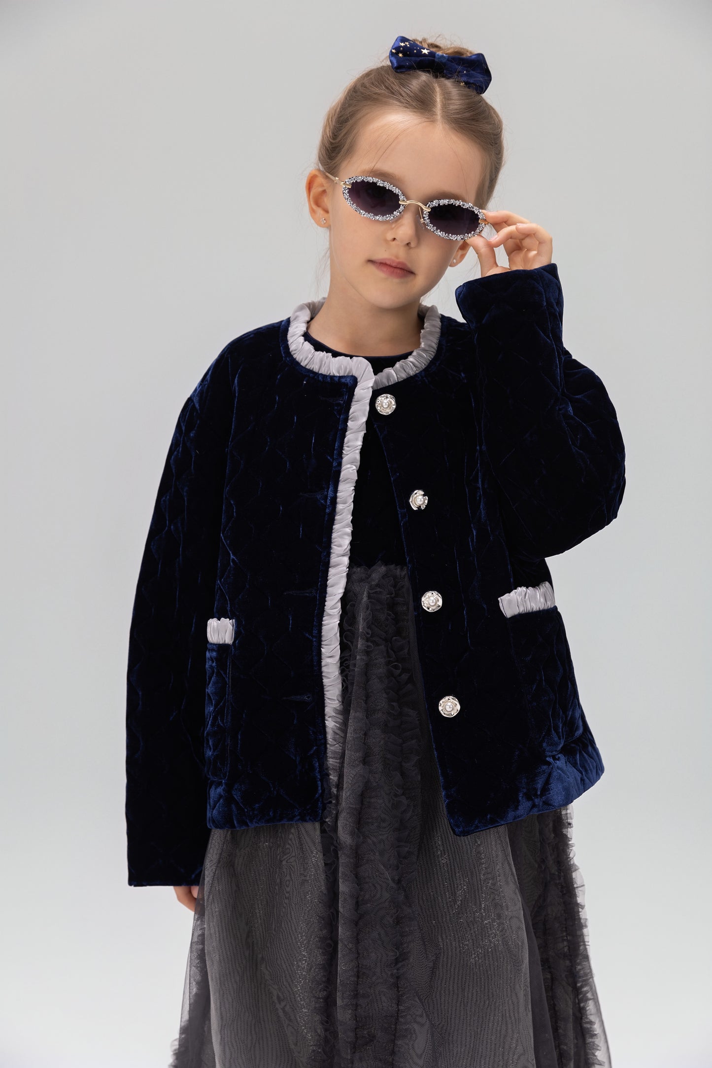 A young girl wearing sunglasses, a gray dress, and the KIKI and JOJO Girls Quilted Ruffle Jacket in sapphire blue stands against a plain background.