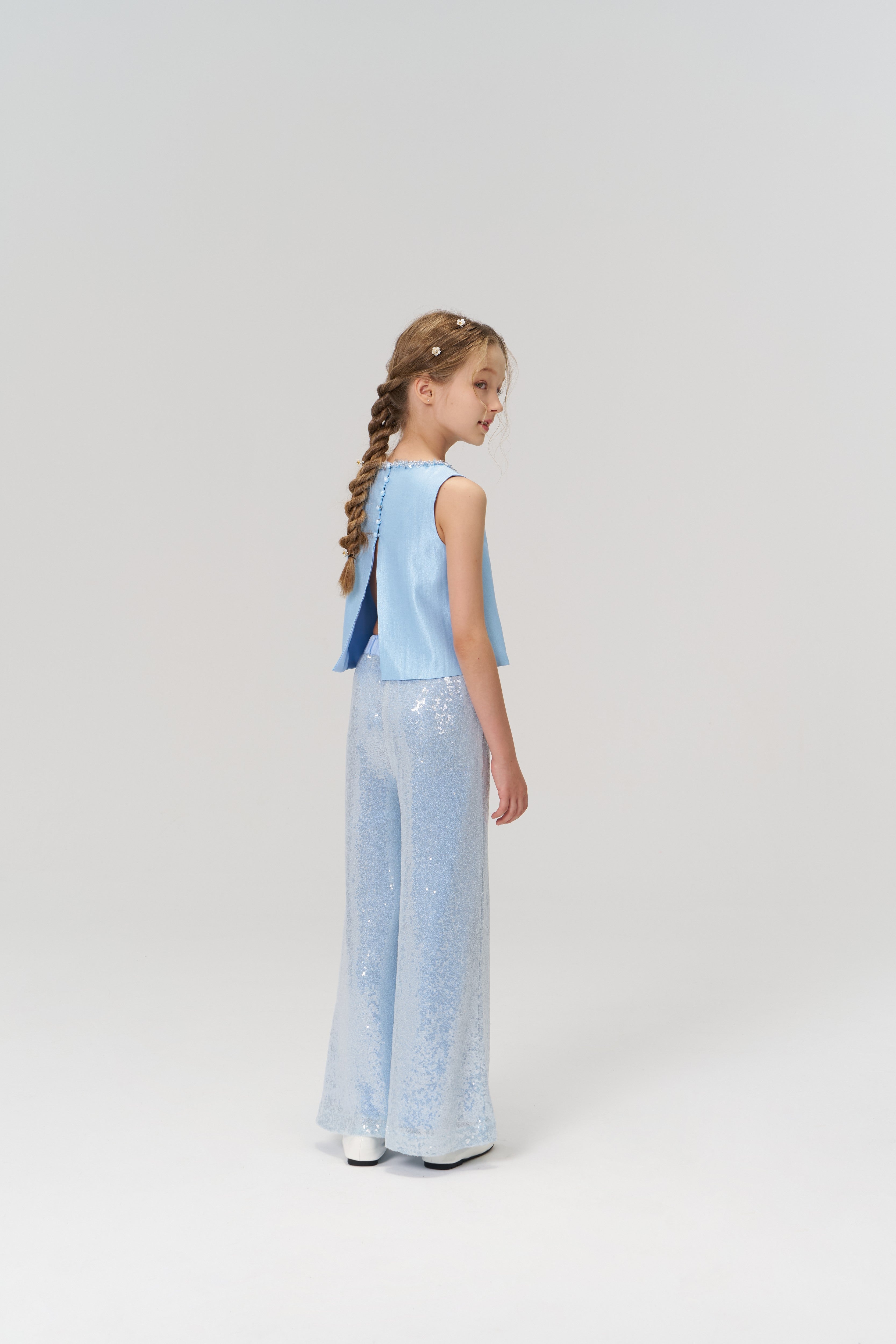 A young girl with braided hair stands facing away, wearing a light blue sleeveless top and KIKI and JOJO's Radiance Sequin Wide-leg Pants against a plain light background.