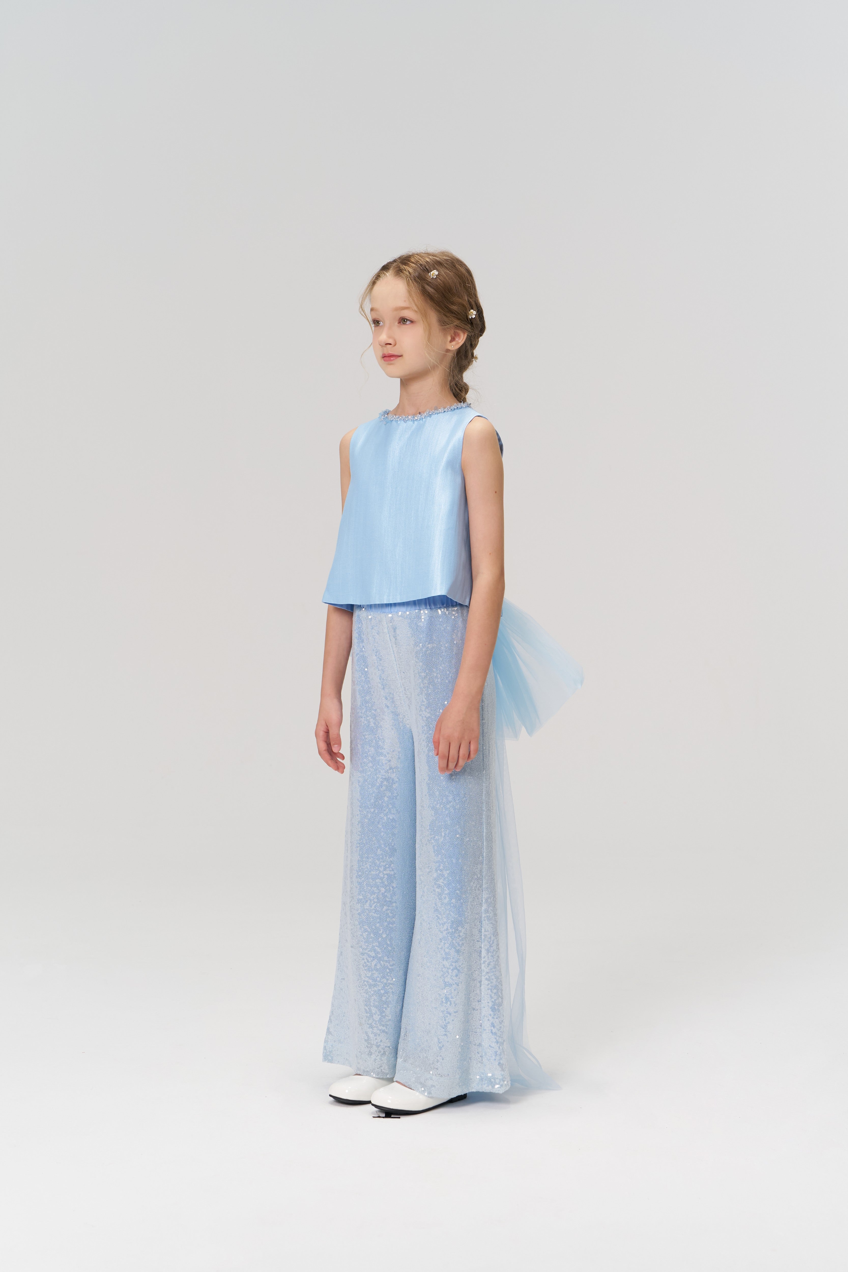 A young girl poses in a studio wearing KIKI and JOJO’s Radiance Sequin Wide-leg Pants, a sleeveless light blue top, and white shoes, all set against a simple light-colored backdrop.