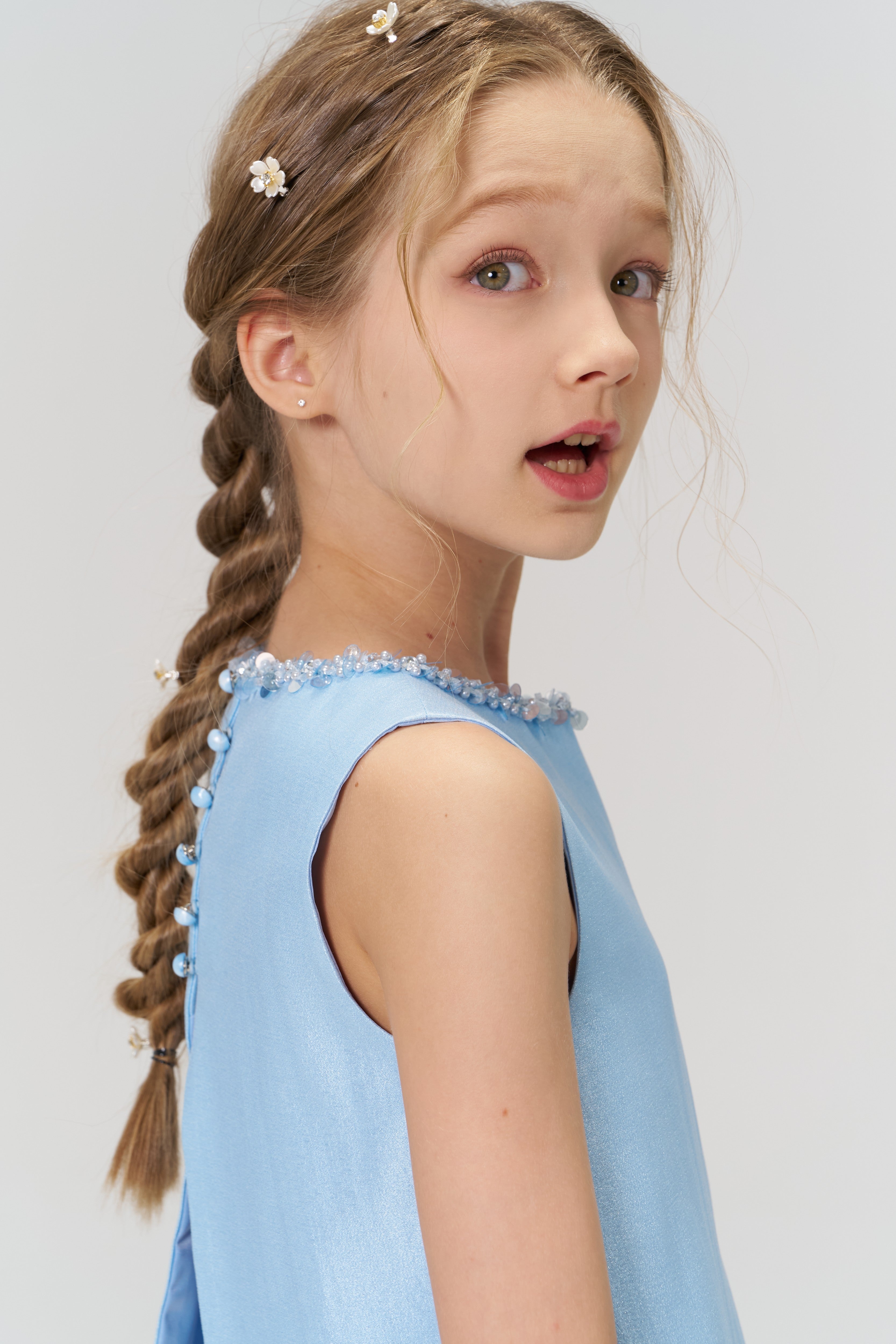 A young girl with braided hair and floral hairpins wears the KIKI and JOJO Radiance Satin Crop Top in light blue, looking slightly over her shoulder—a stunning pick for holiday parties.