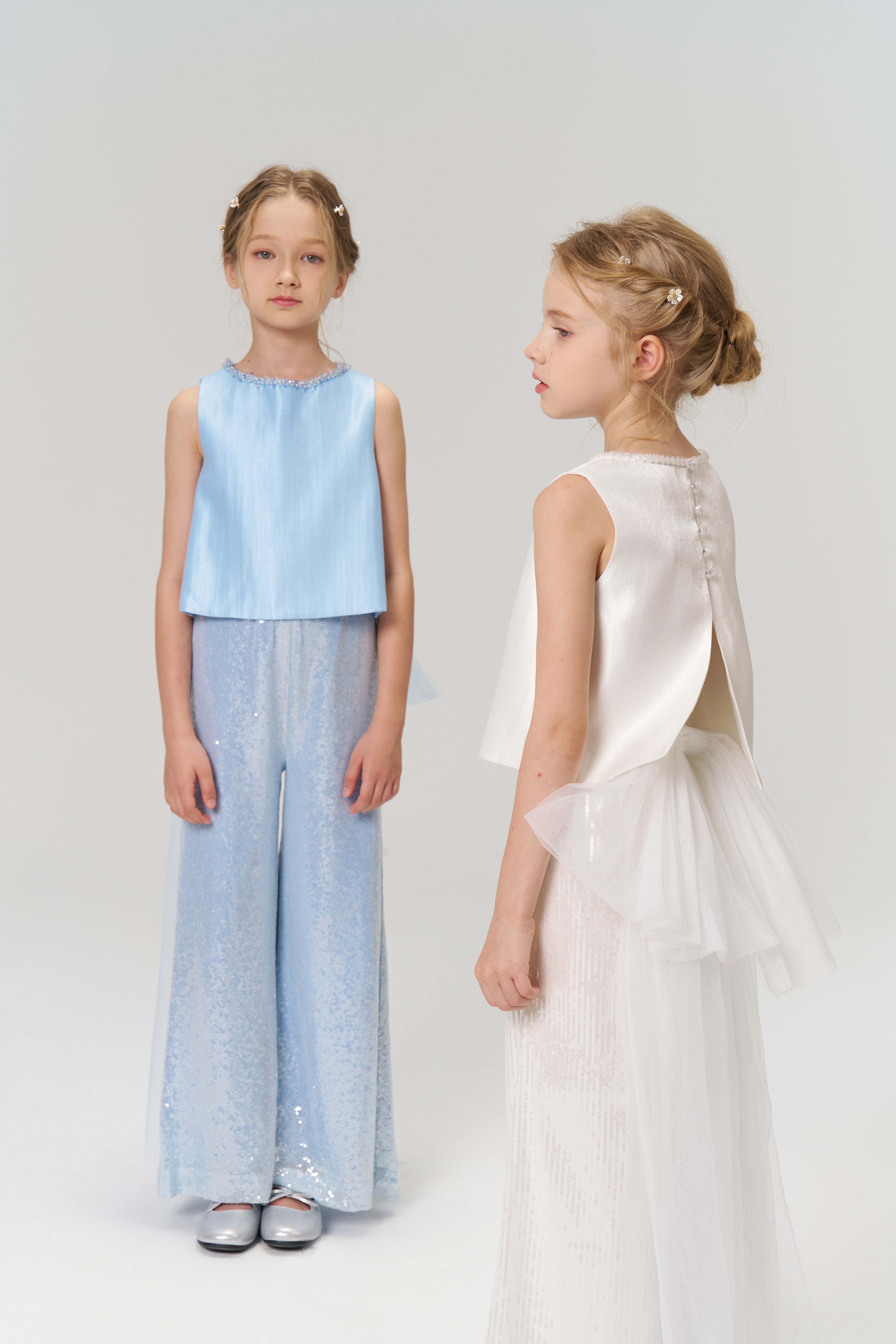 Two young girls stand in a studio; one wears the KIKI and JOJO Radiance Sequin Wide-Leg Pants with a light blue sleeveless top, while the other wears a white sleeveless top with a detachable tulle bow and matching skirt.