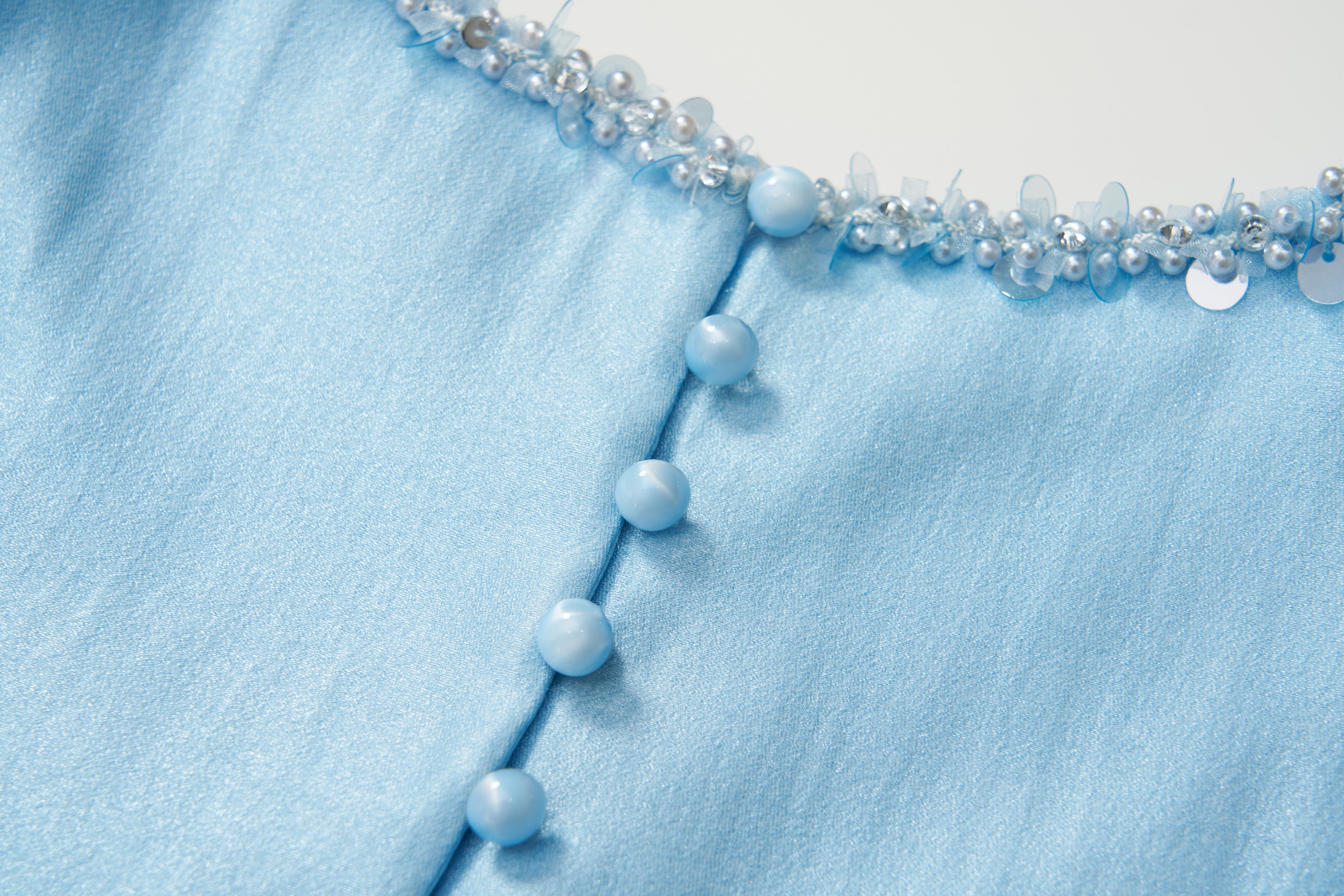 Close-up of the Radiance Satin Crop Top by KIKI and JOJO in light blue with a liquid-sheen finish, round buttons, and a hand-beaded pearl neckline embellished with sequins.