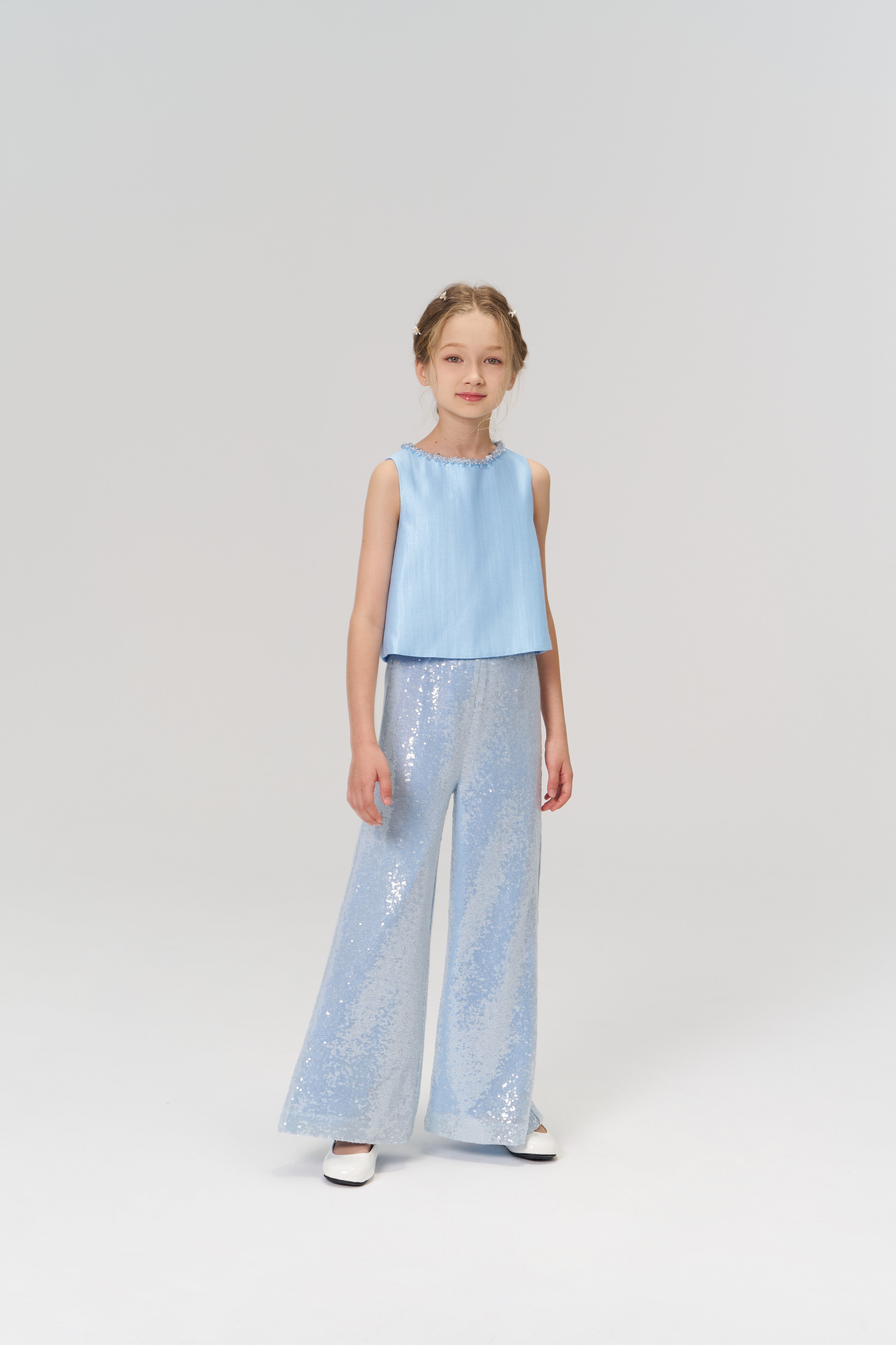 A young girl faces forward, wearing KIKI and JOJO’s Radiance Sequin Wide-leg Pants in light blue with a sleeveless top and white shoes, set against a plain light background.