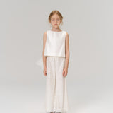 A young girl faces forward in the KIKI and JOJO Radiance Satin Crop Top with a hand-beaded pearl neckline, paired with white sequin wide-leg pants and silver shoes—an ideal look for holiday parties against a plain background.