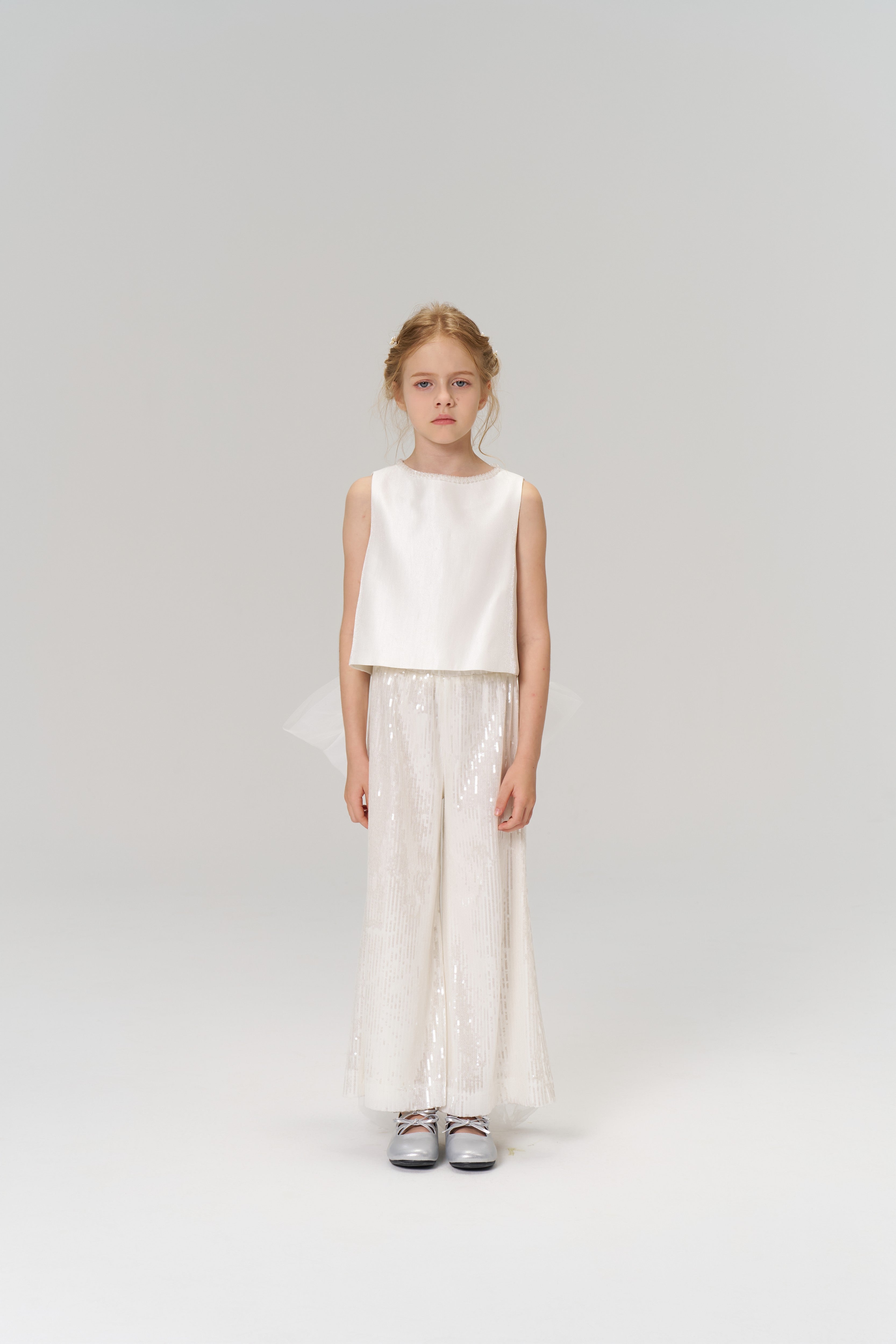 A young girl faces forward in the KIKI and JOJO Radiance Satin Crop Top with a hand-beaded pearl neckline, paired with white sequin wide-leg pants and silver shoes—an ideal look for holiday parties against a plain background.